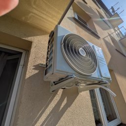 AllinClima Marek Gąsiorowski - Zewnętrzna jednostka klimatyzacji Rotenso zamontowana na elewacji budynku mieszkalnego, widok z dołu, z fragmentem okna i czyimś palcem w kadrze.