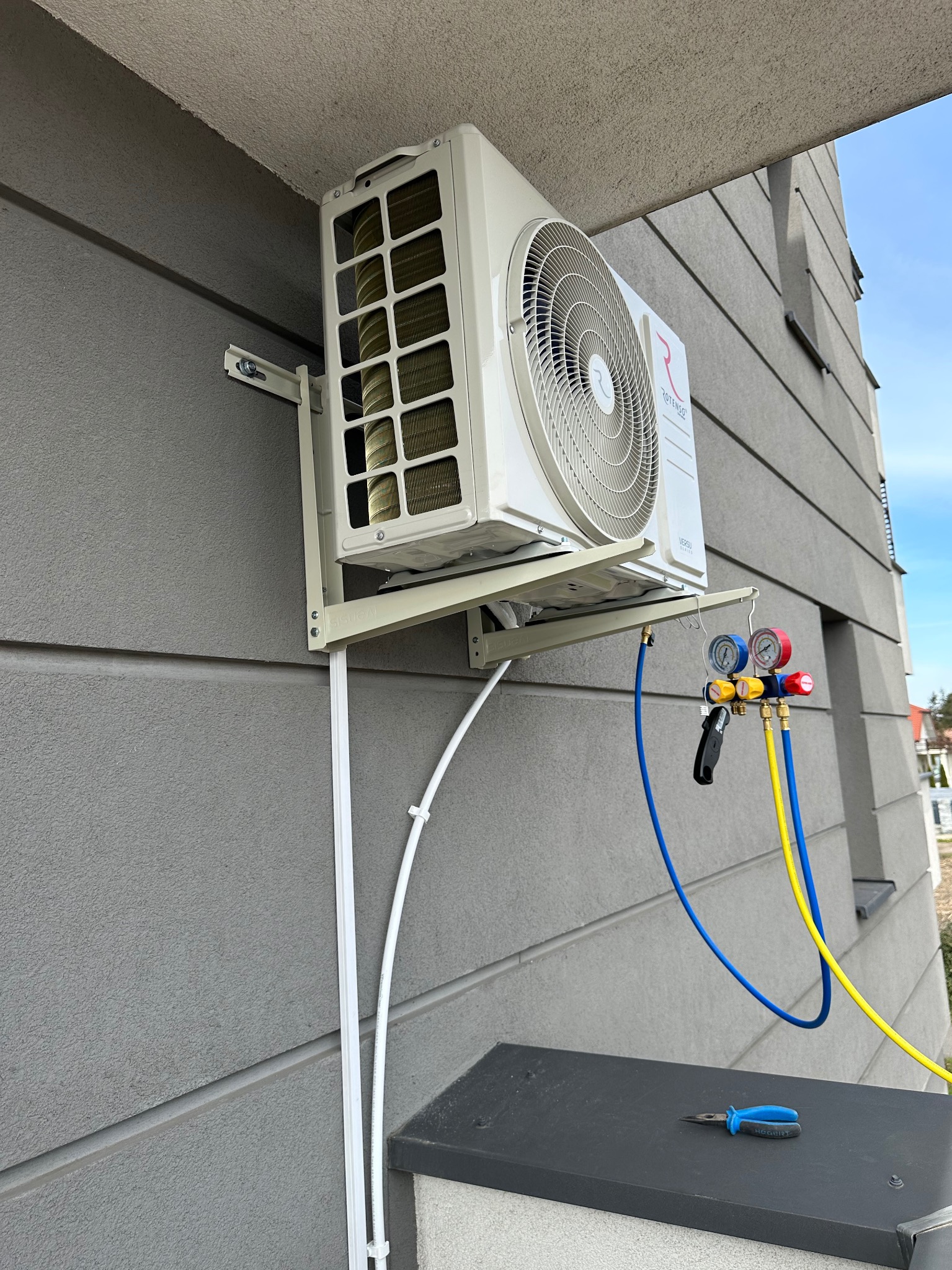 Biały klimatyzator na szarej elewacji budynku, podłączony do manometrów serwisowych; obok leżą szczypce, widoczne przewody instalacyjne.