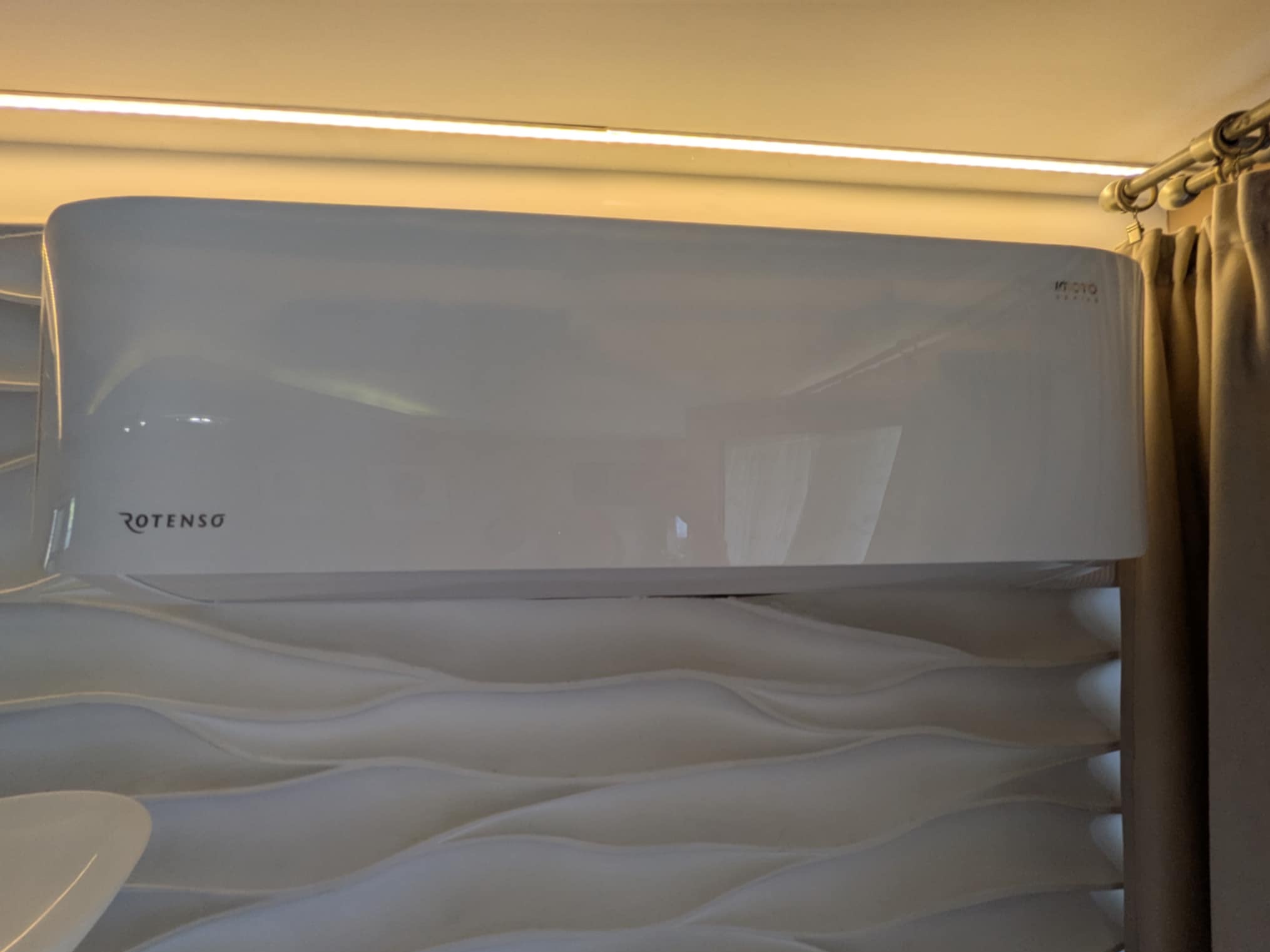 Biała klimatyzacja Rotenso na ścianie z dekoracyjną fakturą fal, oświetlona listwą LED, z zasłoną po prawej stronie kadru. Widoczne logo marki.