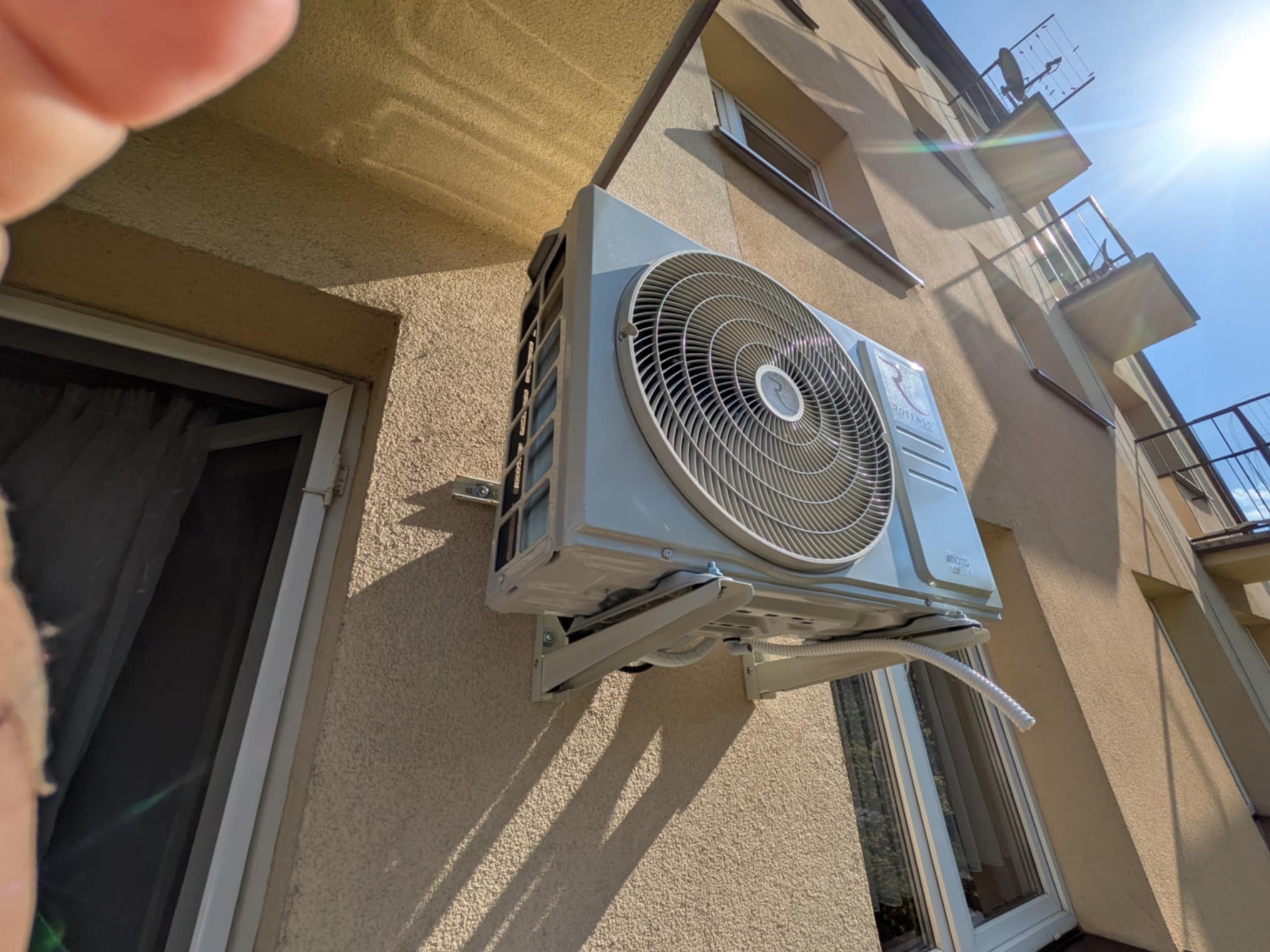 Zewnętrzna jednostka klimatyzacji Rotenso zamontowana na elewacji budynku mieszkalnego, widok z dołu, z fragmentem okna i czyimś palcem w kadrze.
