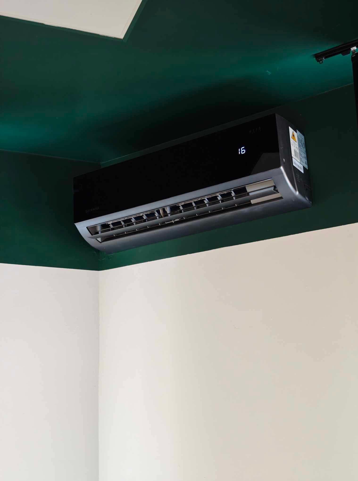Czarny klimatyzator Rotenso z cyfrą 16 na wyświetlaczu, zamontowany na ścianie, gdzie górna część jest zielona, a dolna biała.