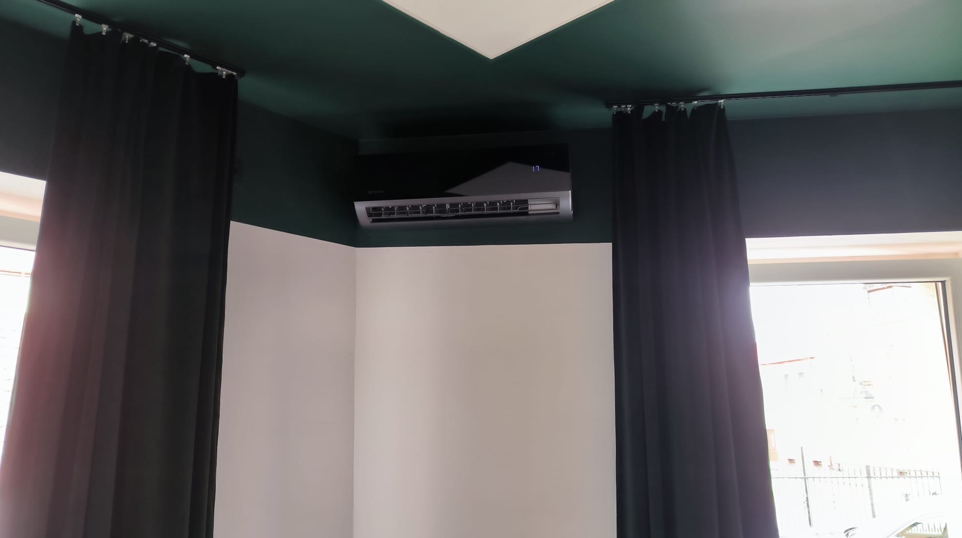 Nowoczesny klimatyzator ścienny w pokoju z ciemnozielonym sufitem i białymi ścianami, zasłonięty częściowo czarnymi zasłonami. Widoczny wyświetlacz z temperaturą.