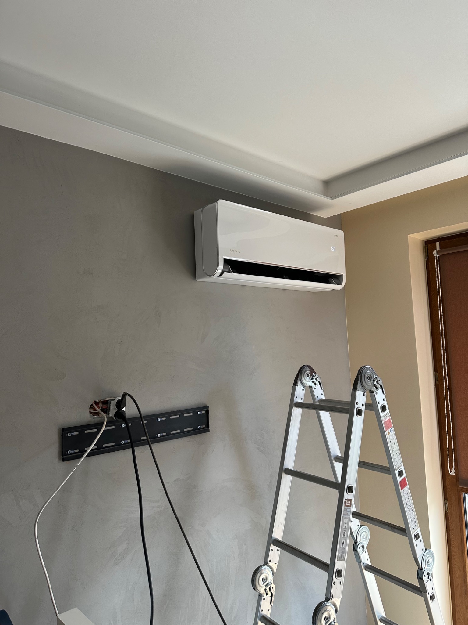 Biała klimatyzacja ścienna zamontowana nad uchwytem TV, z widoczną drabiną, przewody i gładź na ścianie. Montaż klimatyzacji w domu.