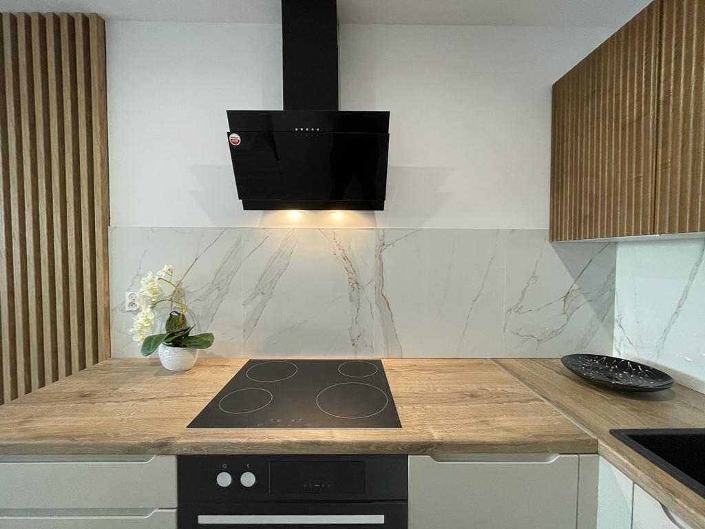 Nowoczesna kuchnia z drewnianymi akcentami: okładzina ścienna imitująca marmur, czarny okap i płyta grzewcza. Blat w kolorze jasnego drewna, minimalistyczny design.