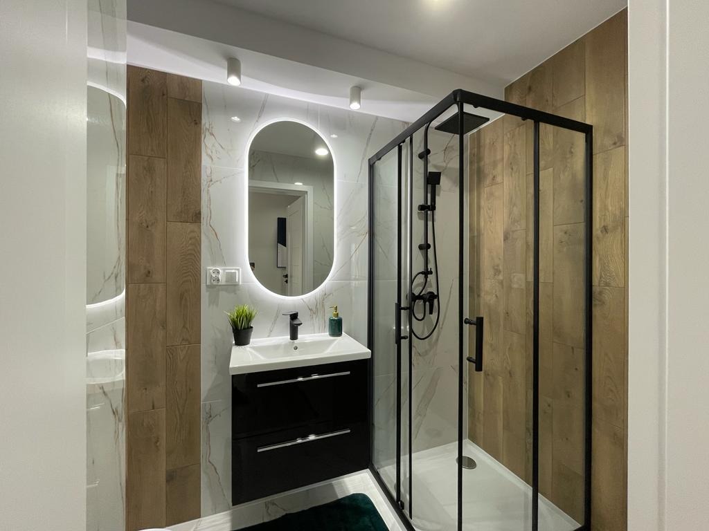 Nowoczesna łazienka z kabiną prysznicową typu walk-in z czarnymi profilami, owalnym lustrem z podświetleniem LED i umywalką z szafką w kolorze czarnym.