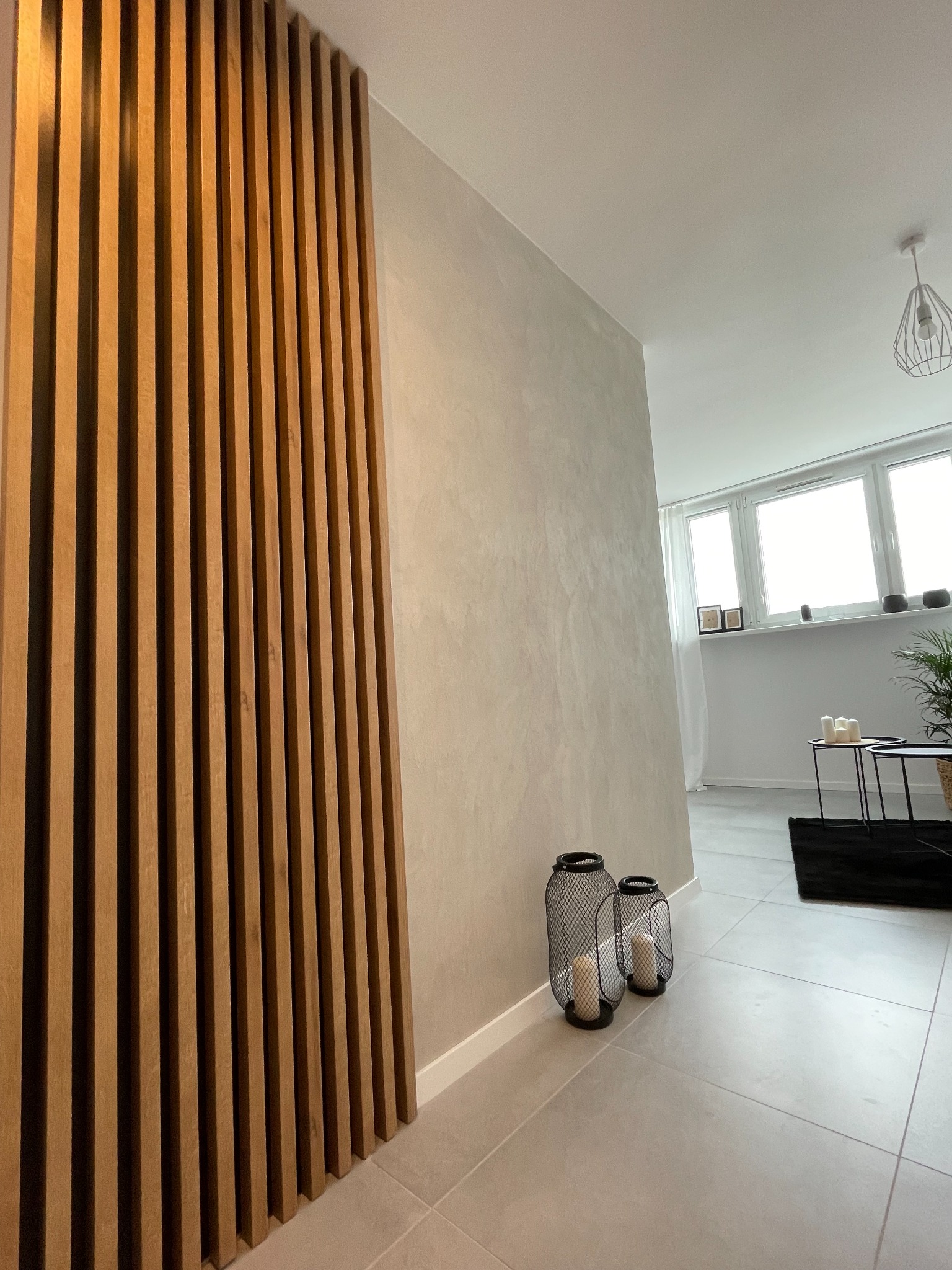 Nowoczesne wykończenie wnętrza: drewniane lamele na ścianie, dekoracyjna faktura ściany, szare płytki podłogowe i lampiony ze świecami.