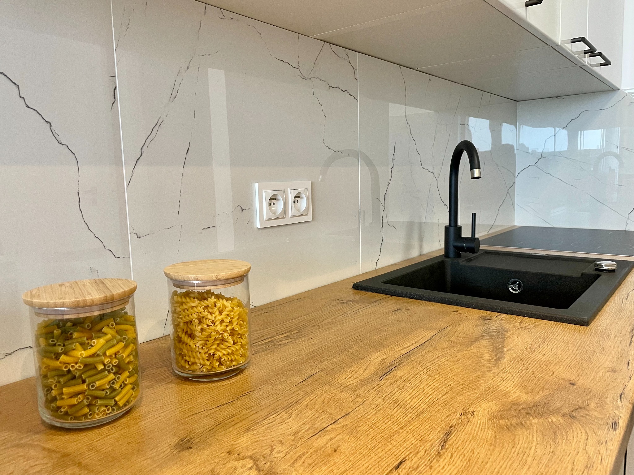 Kuchnia po remoncie: drewniany blat, czarny zlew i bateria, marmurowe płytki na ścianie, gniazdko elektryczne i słoiki z makaronem. Nowoczesny design.