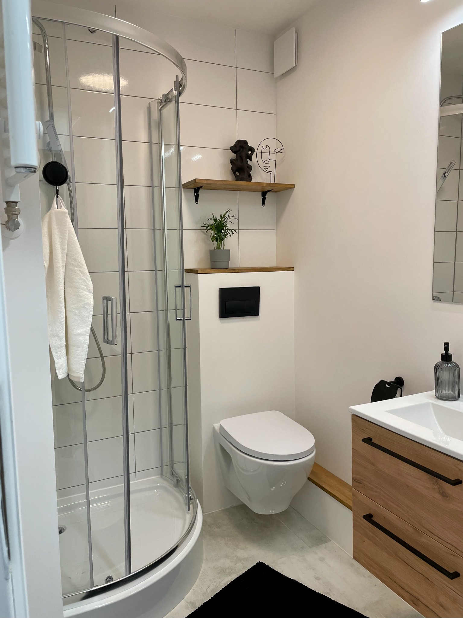 Nowoczesna łazienka z kabiną prysznicową, wiszącą toaletą i drewnianą szafką umywalkową. Minimalistyczny wystrój z akcentami dekoracyjnymi na półce.