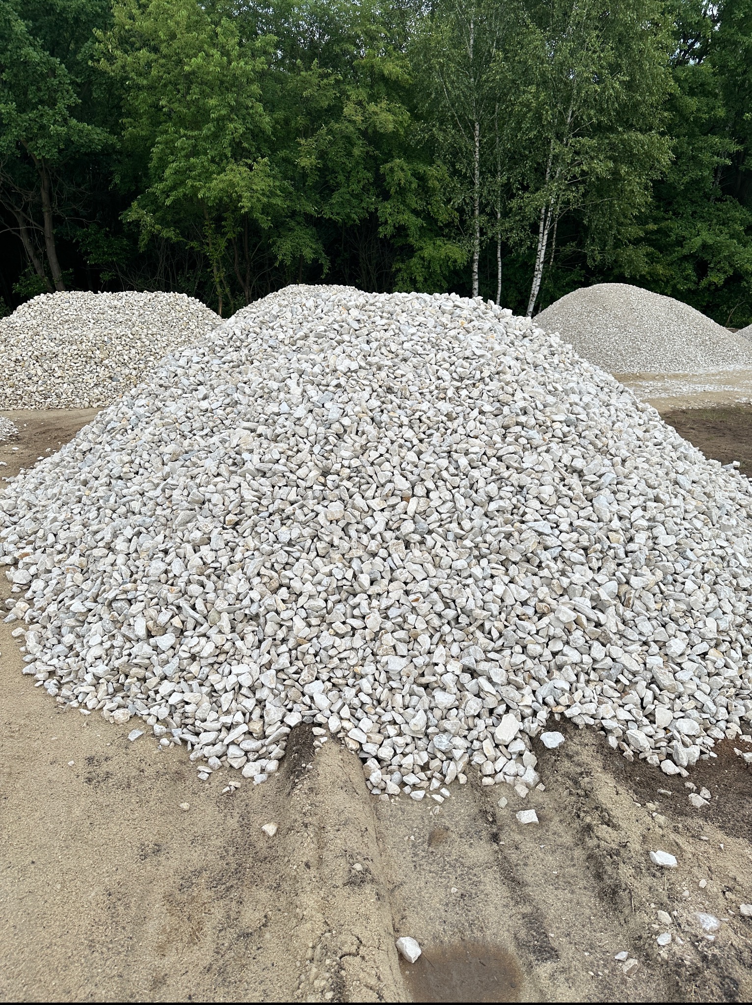 Białe kruszywo na hałdzie ziemi, w tle drzewa. Widoczne ślady opon na ziemi. Materiał sypki przygotowany do transportu lub dalszych prac ziemnych.