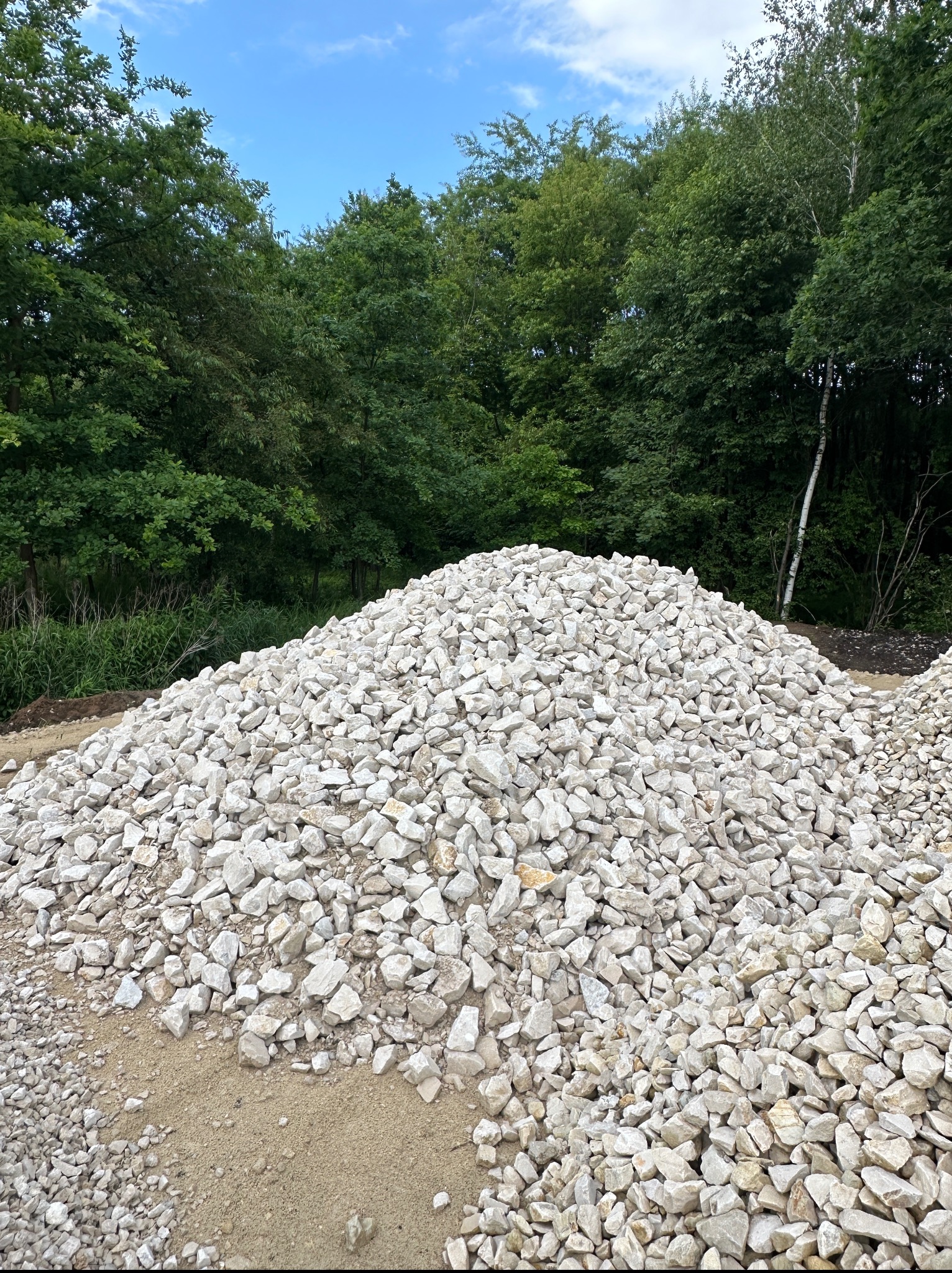 Kupa białego kruszywa na piasku, z zielonym tłem drzew i błękitnym niebem. Kruszywo o nieregularnych kształtach, różne wielkości kamieni.