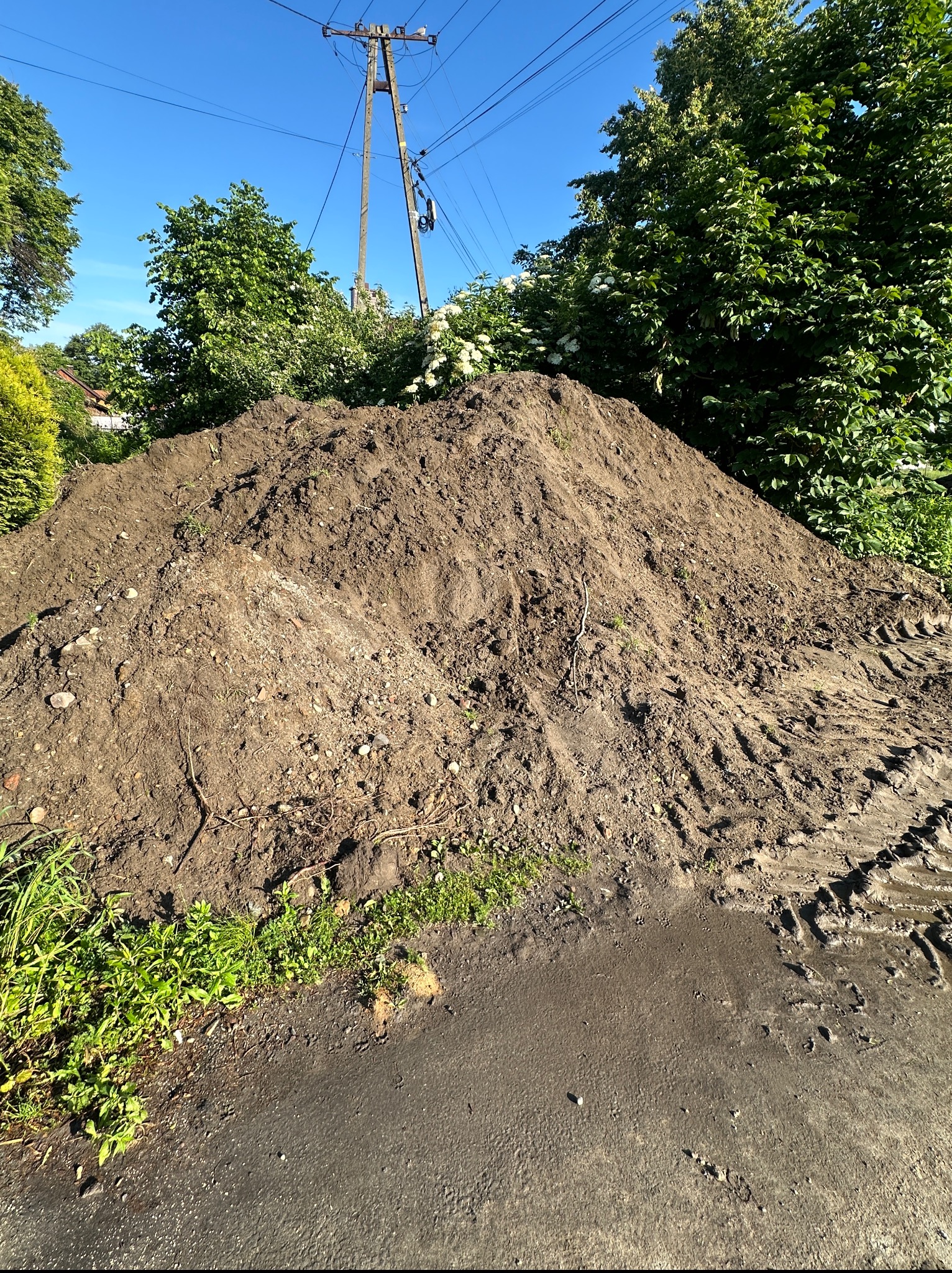 Kupa ziemi z widocznymi śladami opon, zieleń w tle i słup energetyczny pod błękitnym niebem. Roboty ziemne w słoneczny dzień.