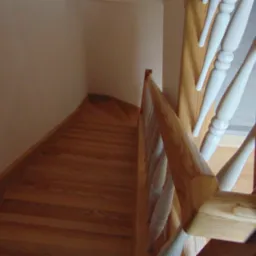 Kątowe ujęcie z góry na dębowe schody z balustradą o jasnych, toczonych tralkach. Widoczne zakręcające stopnie i fragment ściany w neutralnym odcieniu.