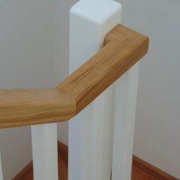 Stolarstwo Szudera Piotr Szudera - Fragment drewnianej balustrady schodowej z jasnego drewna i białymi słupkami, widok z góry, ukazujący detale połączeń elementów.