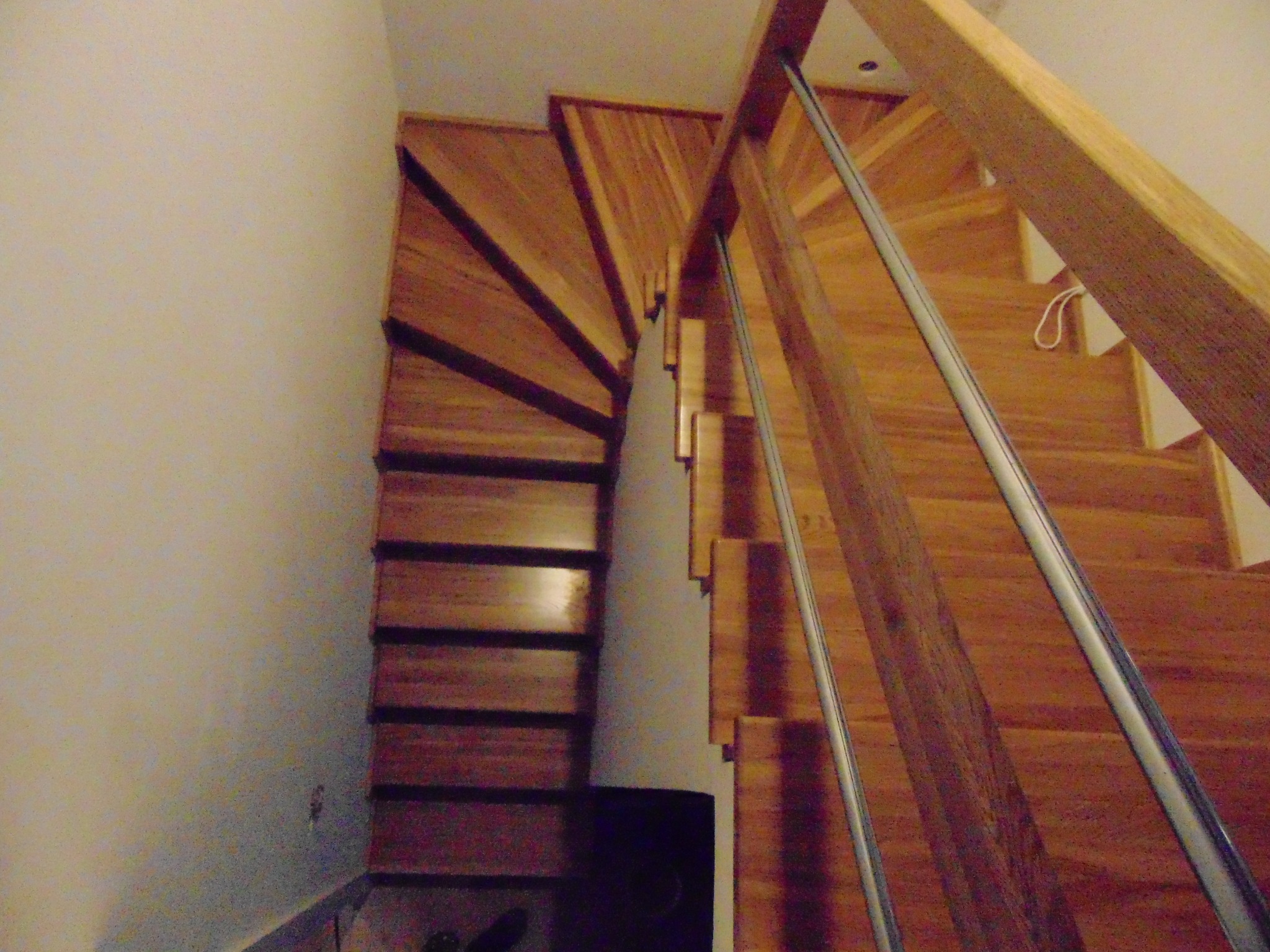 Kątowe ujęcie drewnianych schodów z metalową balustradą, widok z góry. Naturalne oświetlenie podkreśla rysunek drewna. Schody skręcają w lewo, tworząc ciekawy układ.