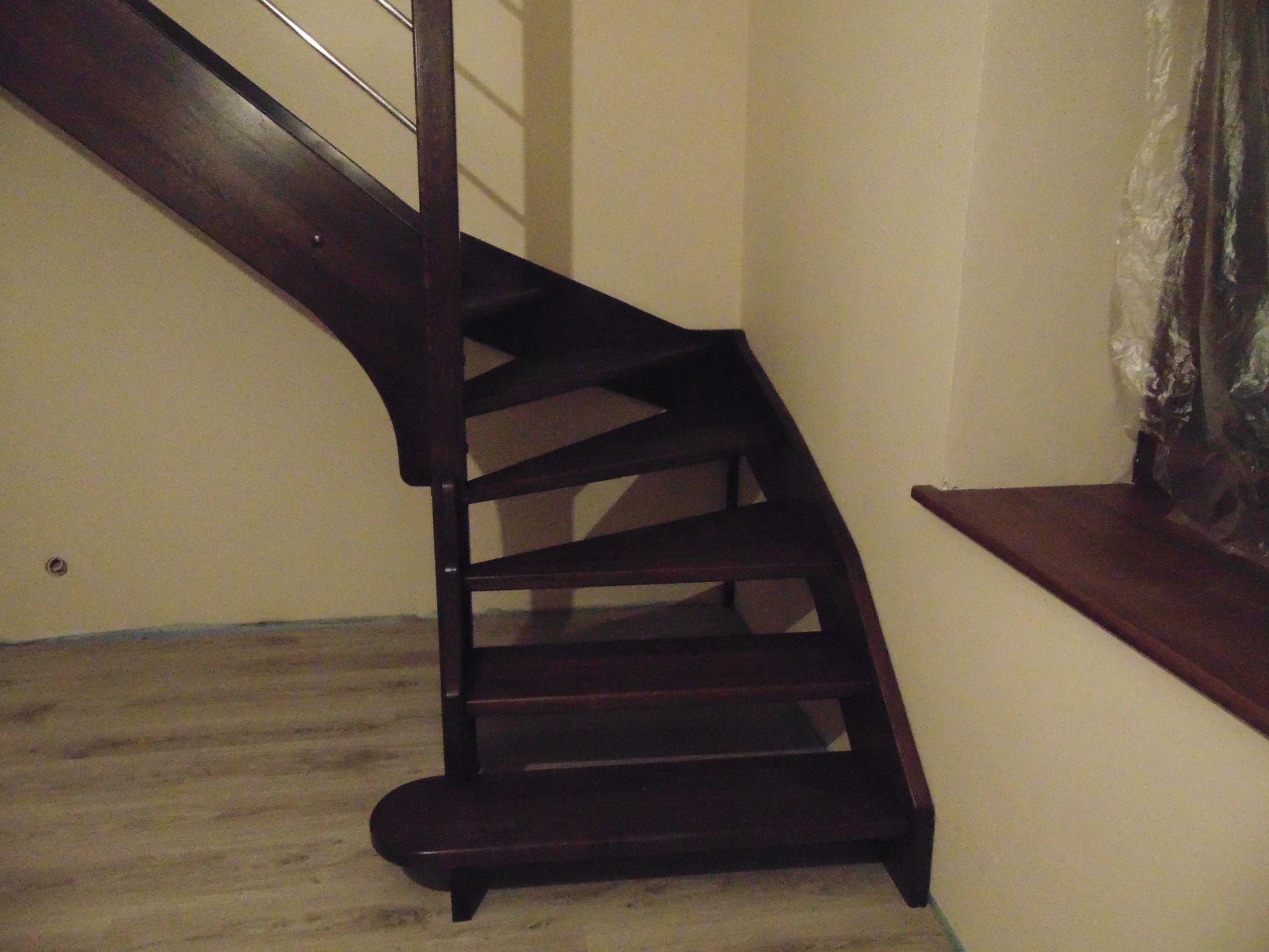 Ciemnobrązowe schody z metalową balustradą, skręcające w lewo, z widoczną drewnianą podłogą i fragmentem okna z parapetem w tle.