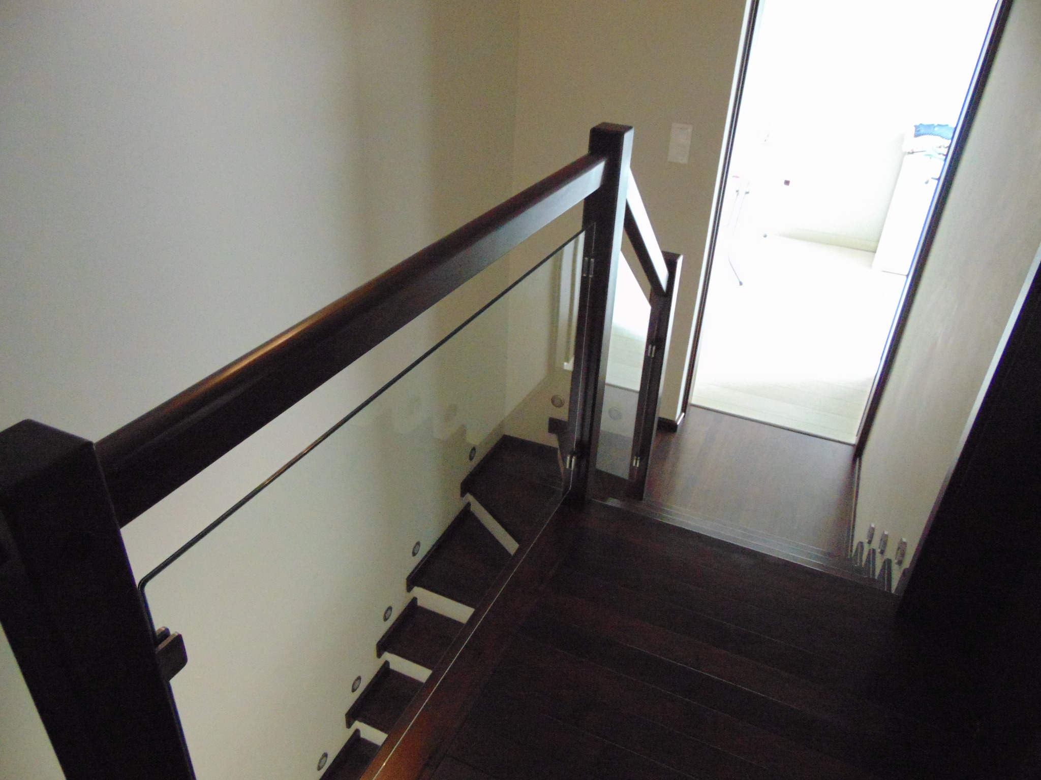 Nowoczesne schody drewniane z ciemnego drewna z szklaną balustradą, oświetleniem punktowym na ścianie i widokiem na jasne pomieszczenie na górze.