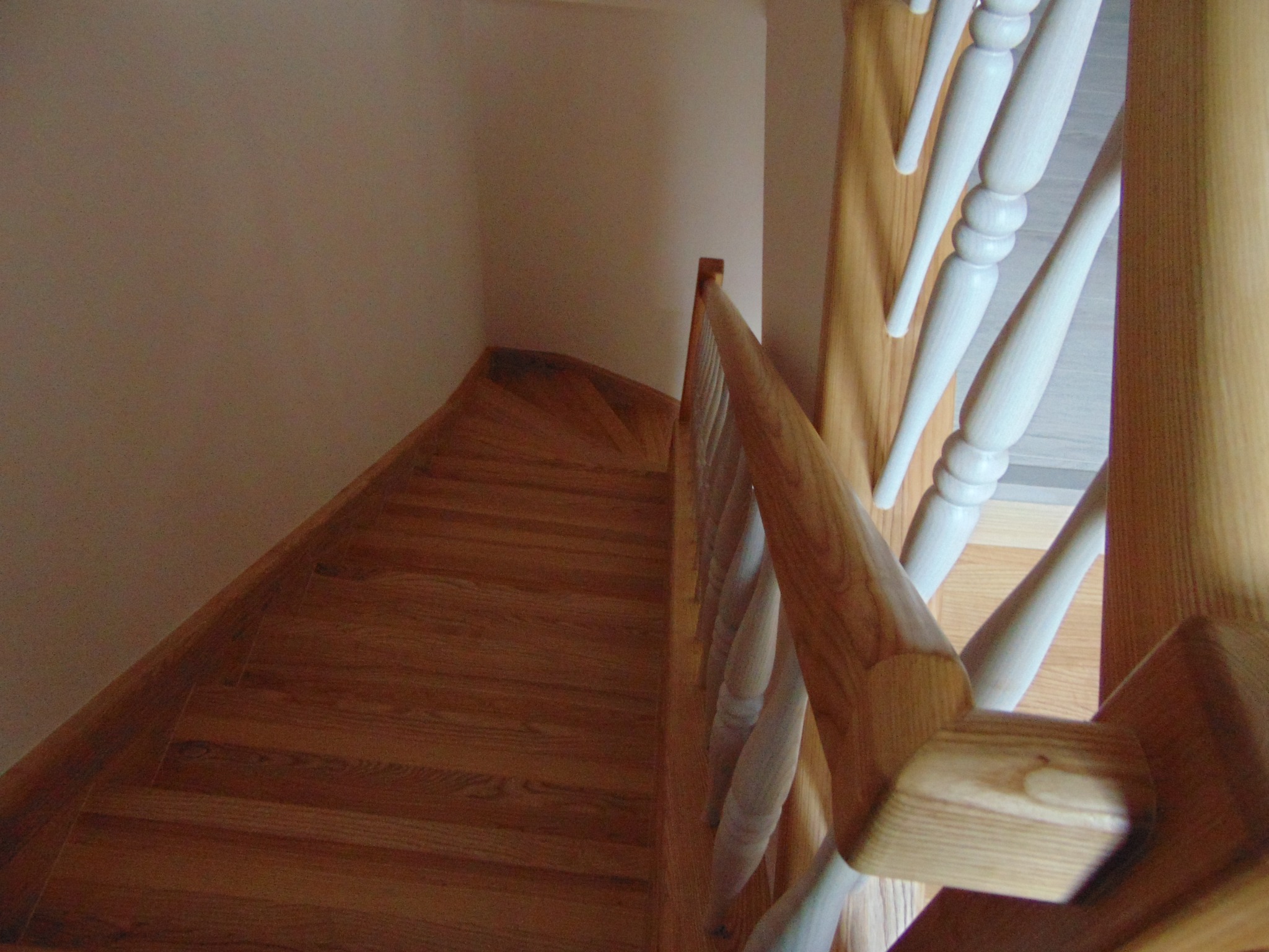Kątowe ujęcie z góry na dębowe schody z balustradą o jasnych, toczonych tralkach. Widoczne zakręcające stopnie i fragment ściany w neutralnym odcieniu.