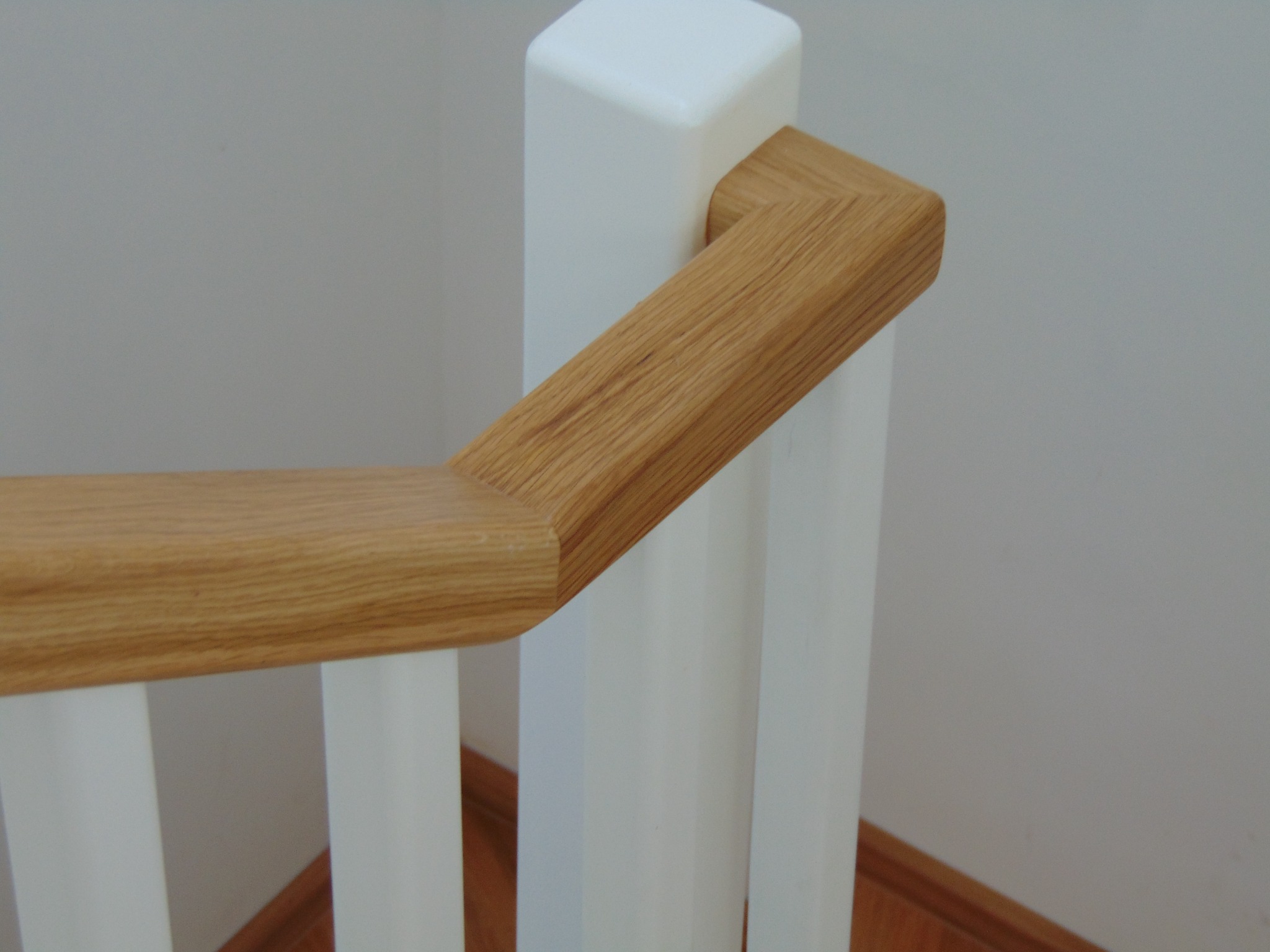 Fragment drewnianej balustrady schodowej z jasnego drewna i białymi słupkami, widok z góry, ukazujący detale połączeń elementów.