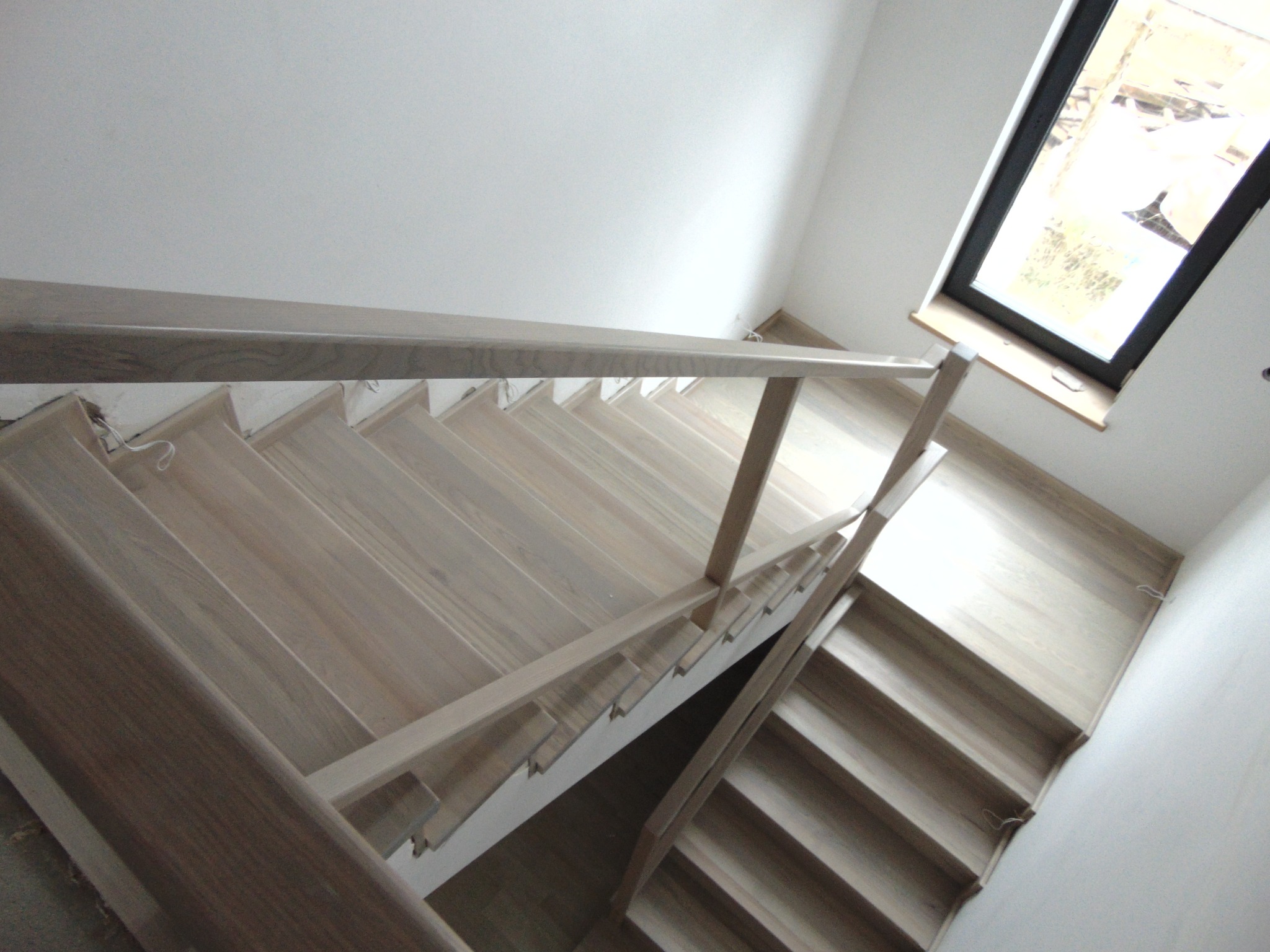 Jasne, minimalistyczne schody z drewna prowadzące do góry, oświetlone przez okno dachowe. Widok z góry, ukazujący strukturę drewna i prostą balustradę.