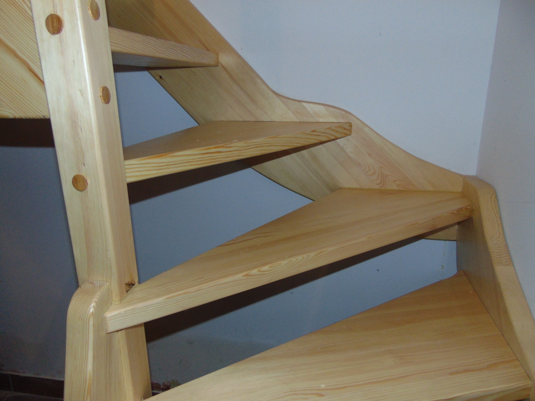 Jasne, drewniane schody z widoczną strukturą drewna, z charakterystycznymi okrągłymi zaślepkami w bocznej belce, ustawione pod kątem na tle jasnej ściany.