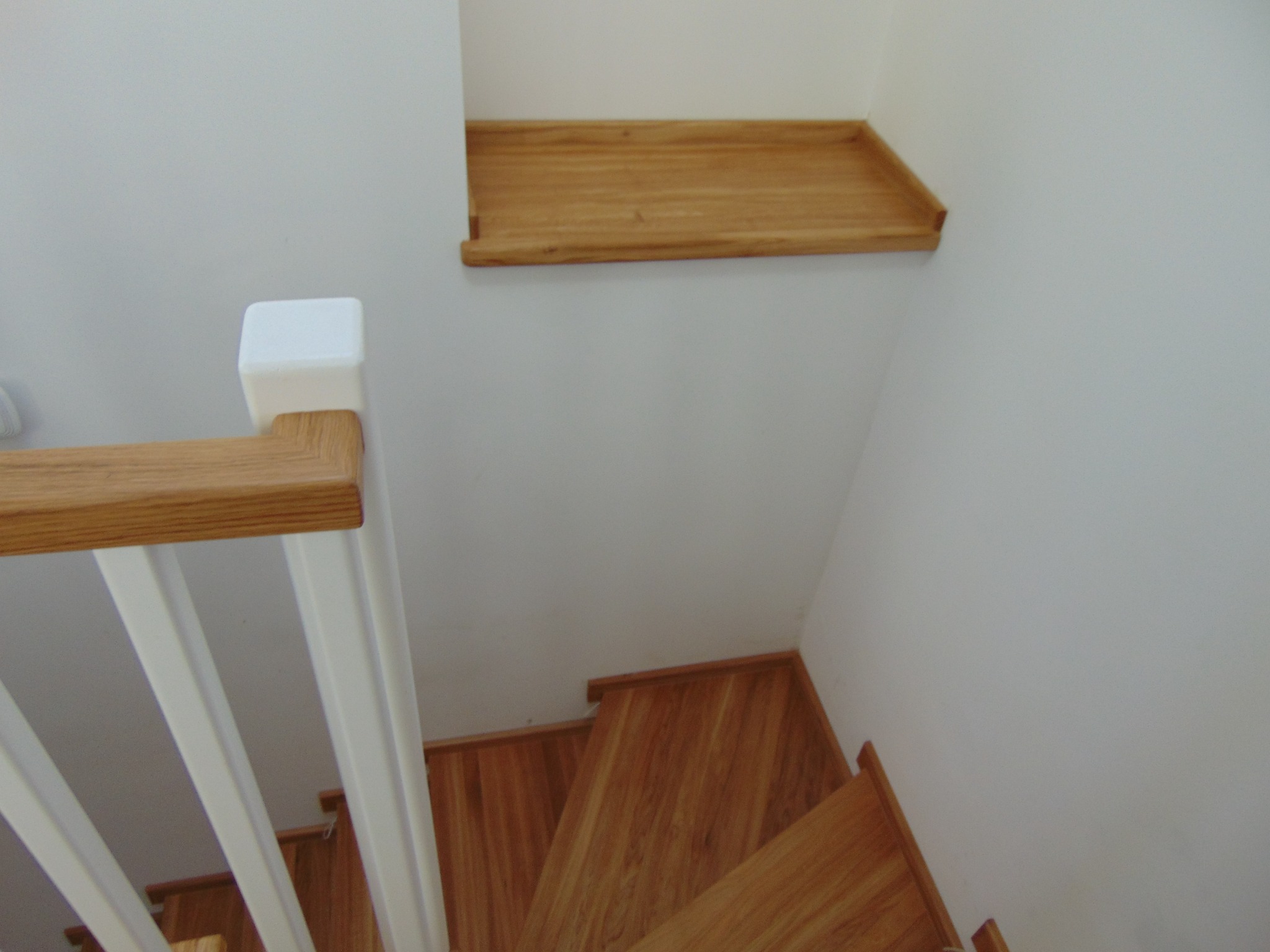 Ujęcie z góry na drewniane schody z białą balustradą. Widoczna półka z drewna na ścianie. Naturalne oświetlenie podkreśla detale wykończenia.