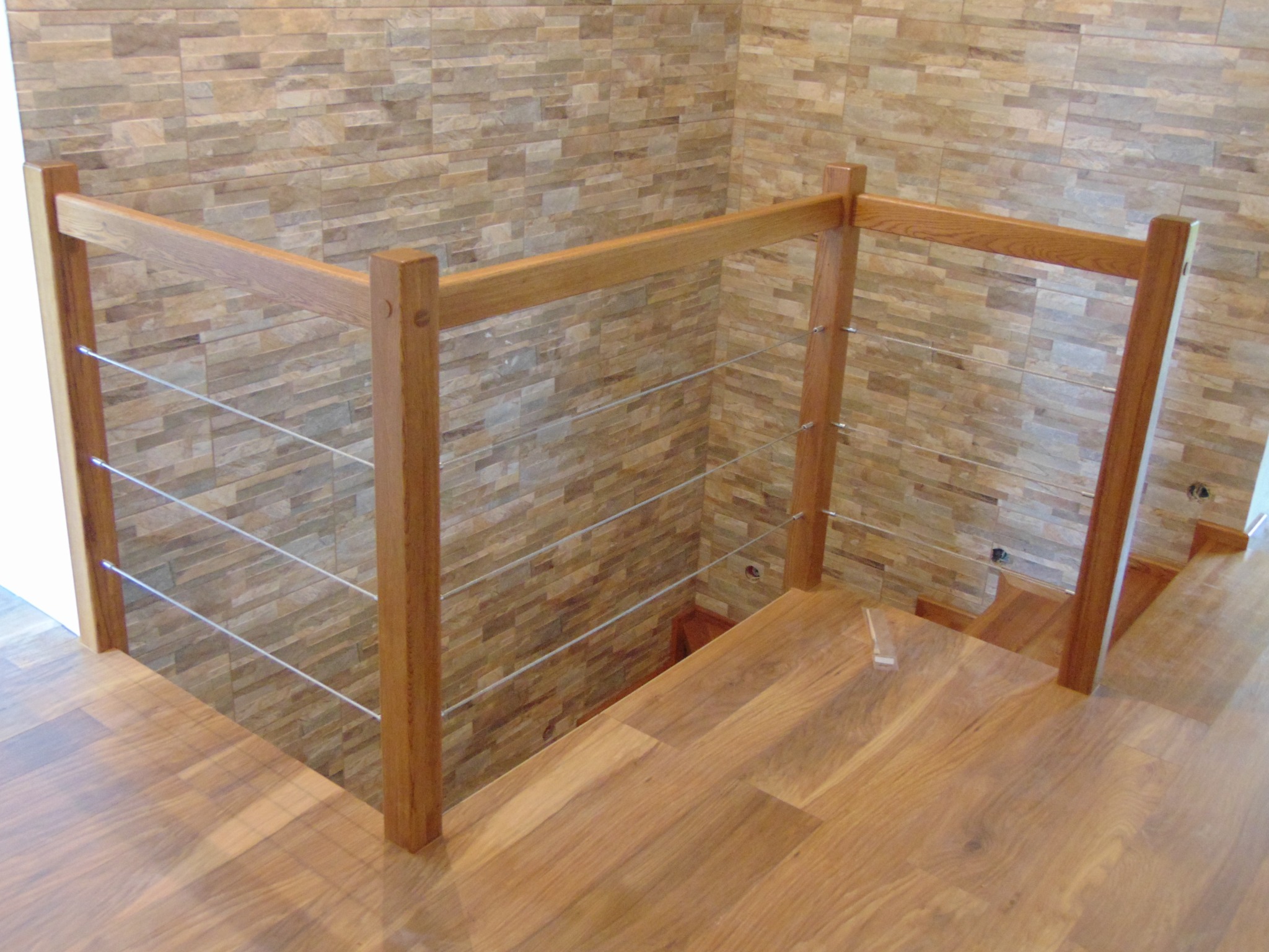 Jasne drewniane schody z balustradą z poziomych linek stalowych, widok z góry na tle ściany z beżowych płytek imitujących kamień.