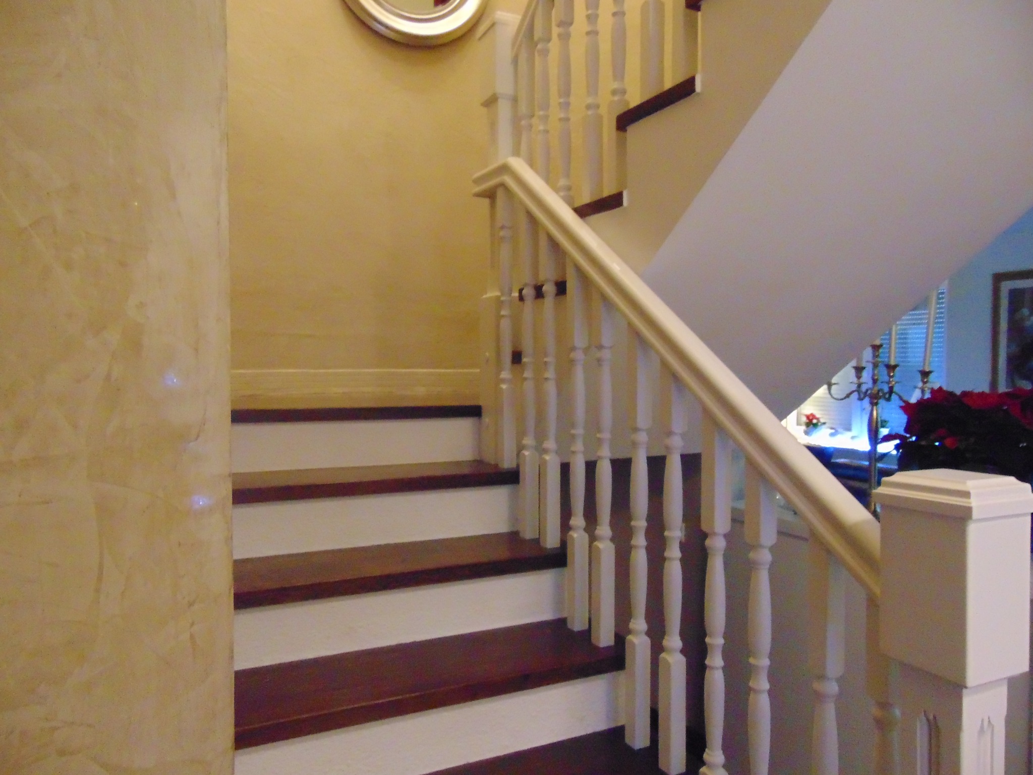 Klasyczne schody z drewnianymi stopniami i białą balustradą, oświetlone ciepłym światłem, widoczne z dołu, z dekoracyjną ścianą w tle.