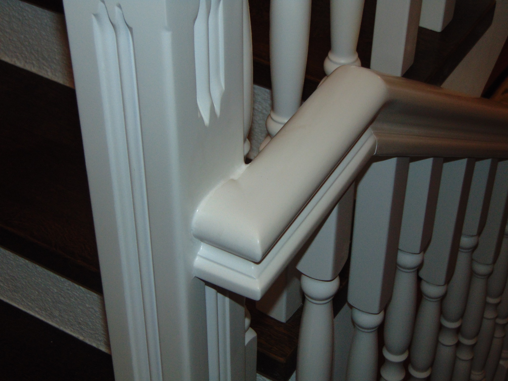 Biała balustrada schodowa z drewnianymi tralkami i ozdobnym słupkiem, widok z bliska na połączenie poręczy i słupka. Nowoczesny design.