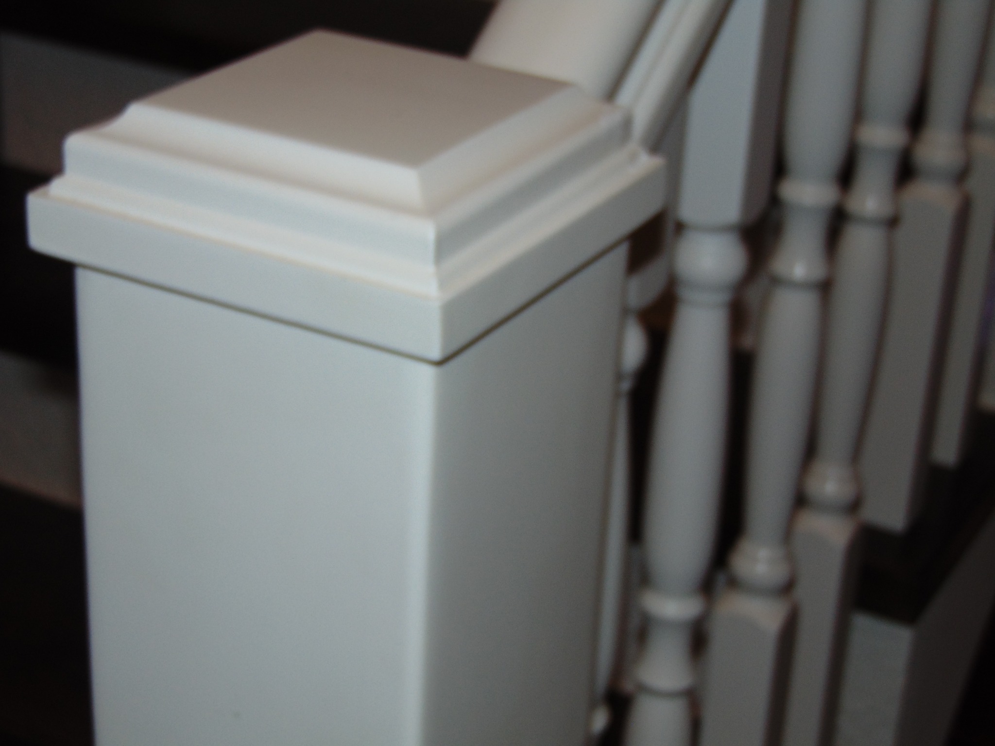 Szczegółowe ujęcie białej, drewnianej balustrady schodowej z dekoracyjnym słupkiem, widoczne stopnie schodów w tle.