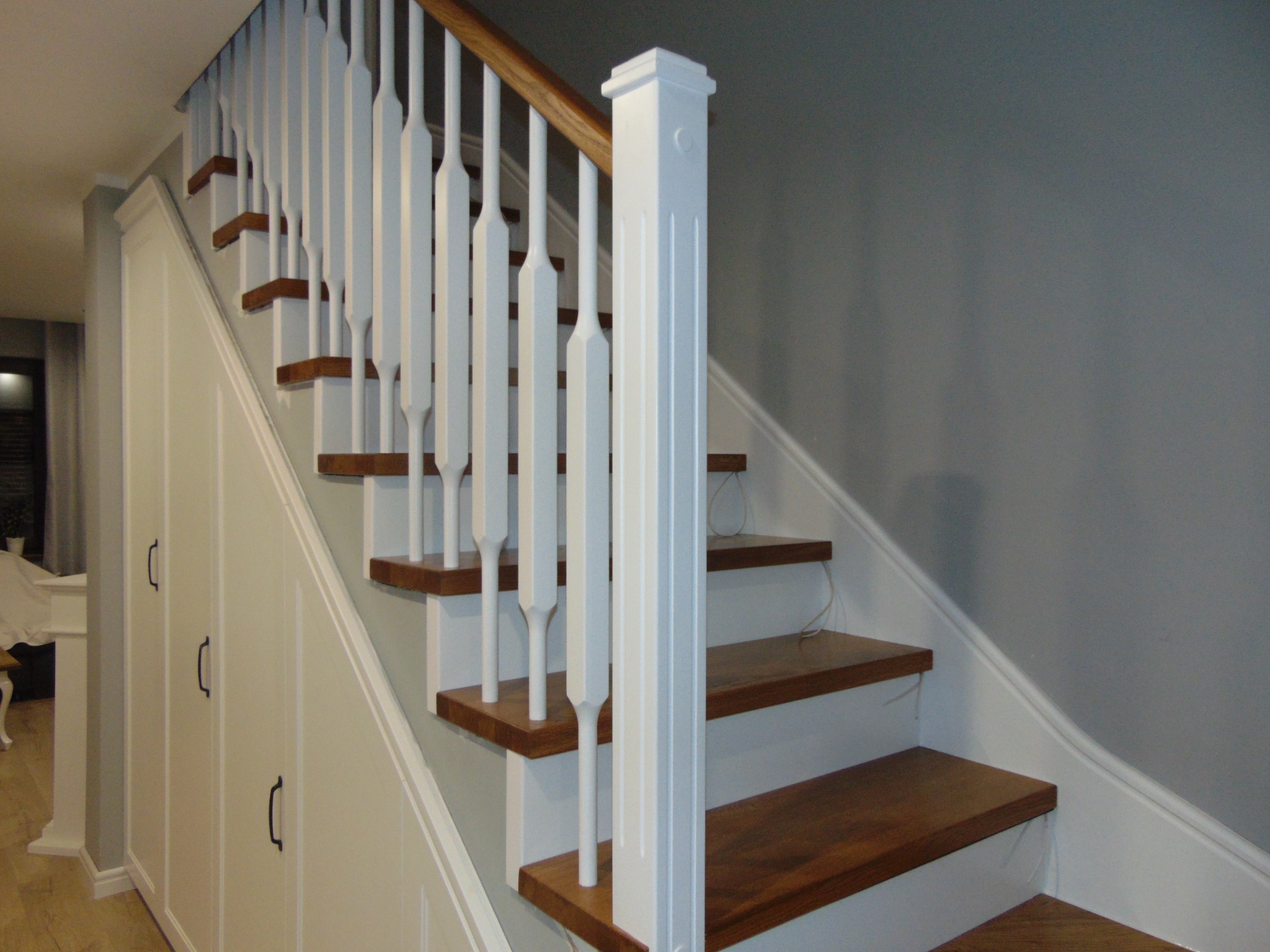 Klasyczne schody z drewnianymi stopniami i białą balustradą w Tarnowie Podgórnym. Pod schodami zabudowa szafkami z ciemnymi uchwytami. Jasne wnętrze.