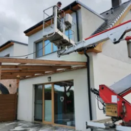 Pracownik na podnośniku czyści elewację jasnego domu jednorodzinnego. Widoczne ślady wody na kostce brukowej, sugerujące czyszczenie. Detal: drewniana pergola.