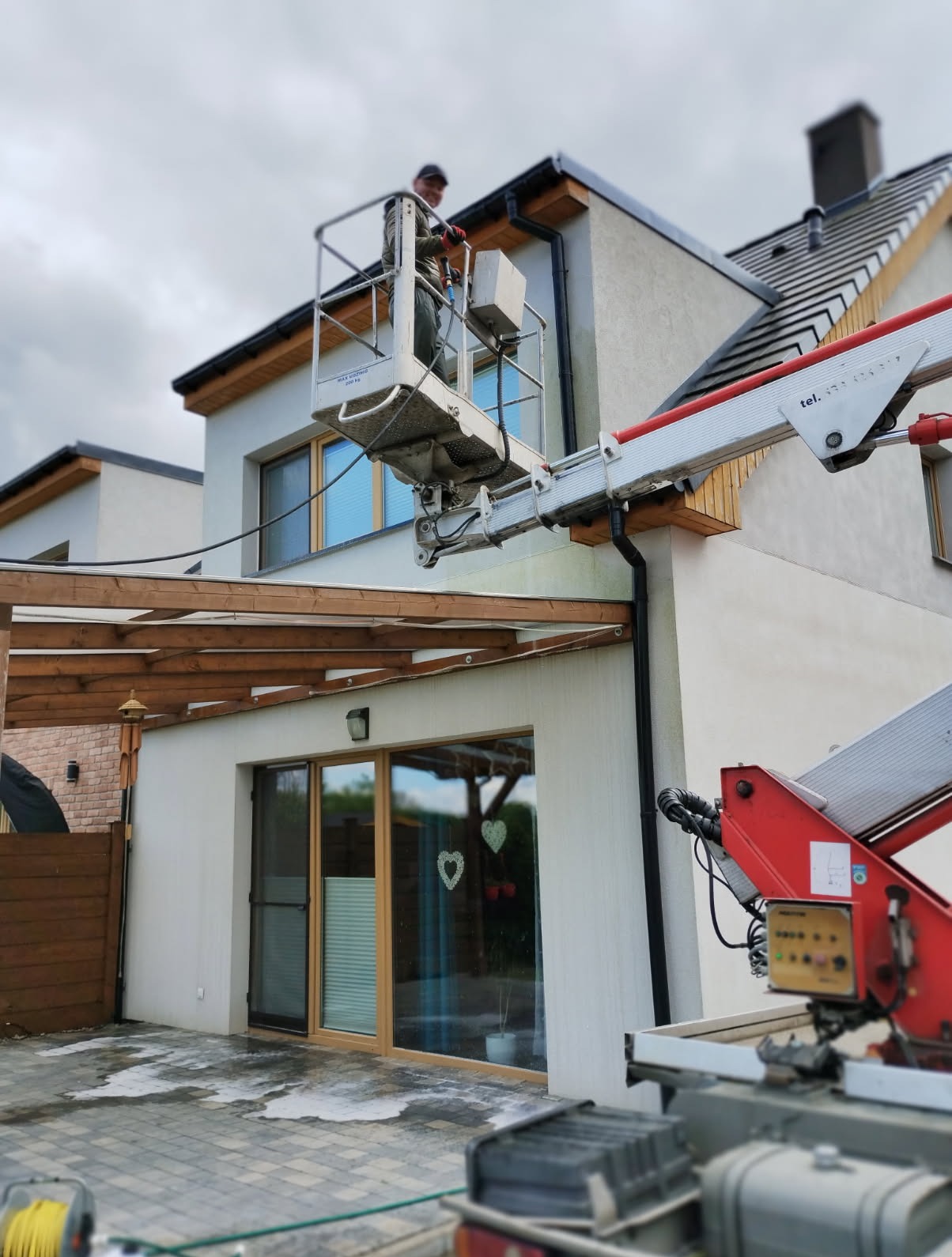 Pracownik na podnośniku czyści elewację jasnego domu jednorodzinnego. Widoczne ślady wody na kostce brukowej, sugerujące czyszczenie. Detal: drewniana pergola.