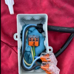 Szara puszka instalacyjna z przewodami i złączkami WAGO 221 na czerwonym tle. Widoczny fragment kabla i kołek rozporowy. Detal instalacji.