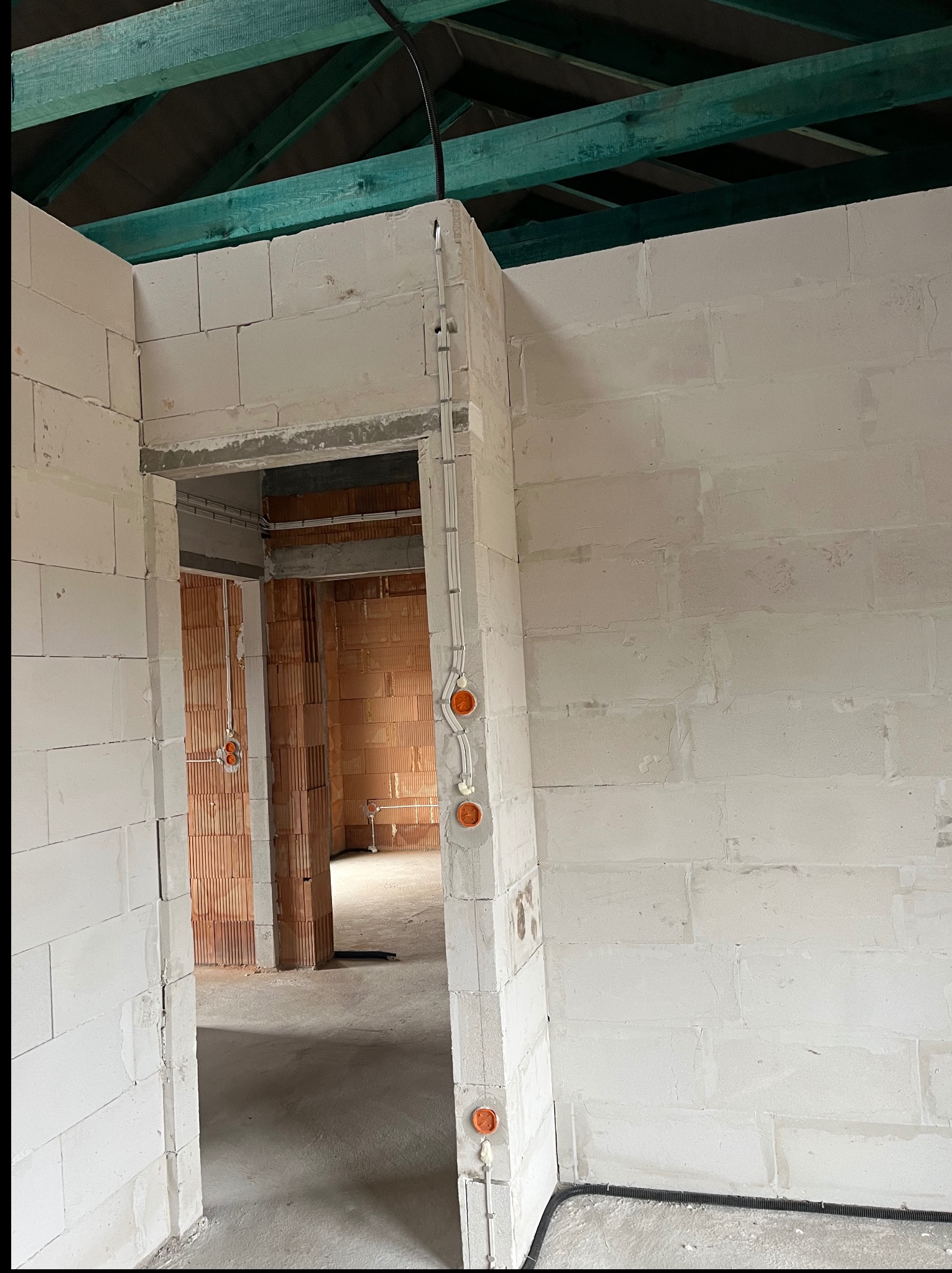Wnętrze w trakcie budowy z widoczną instalacją elektryczną w ścianach z białego bloczka i pomarańczowymi puszkami. Widoczne elementy konstrukcji dachu.