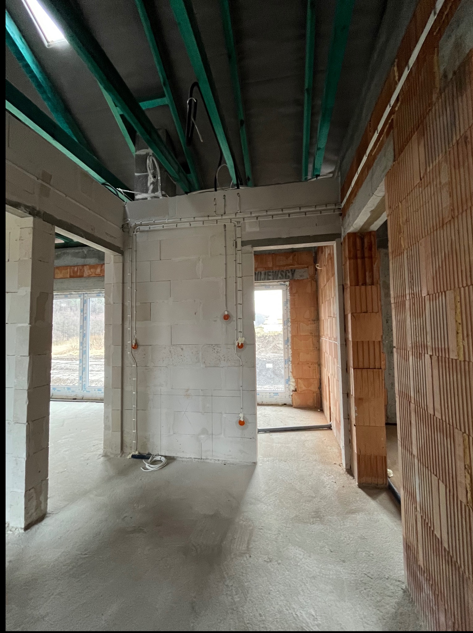 Wnętrze w trakcie budowy z widoczną instalacją elektryczną: kable poprowadzone po ścianach z bloczków, widoczne puszki instalacyjne. Surowy stan, zielone elementy konstrukcji dachu.