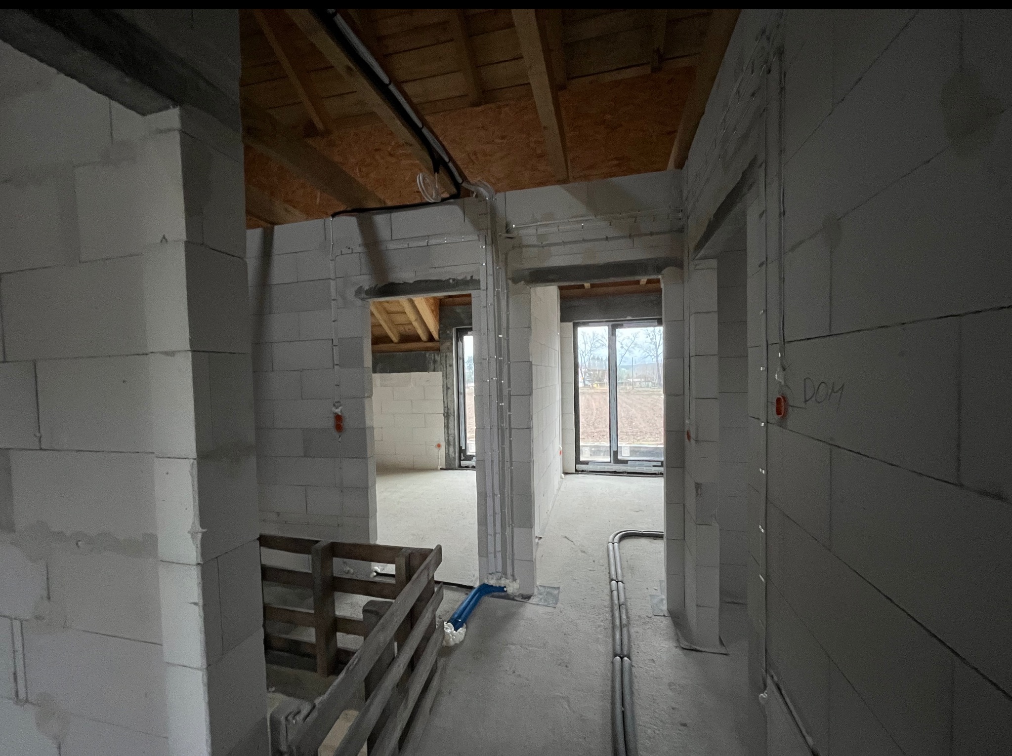 Wnętrze surowego domu w trakcie budowy z widoczną instalacją elektryczną: peszle na ścianach i pod sufitem, okna z widokiem na zewnątrz. Stan surowy zamknięty.