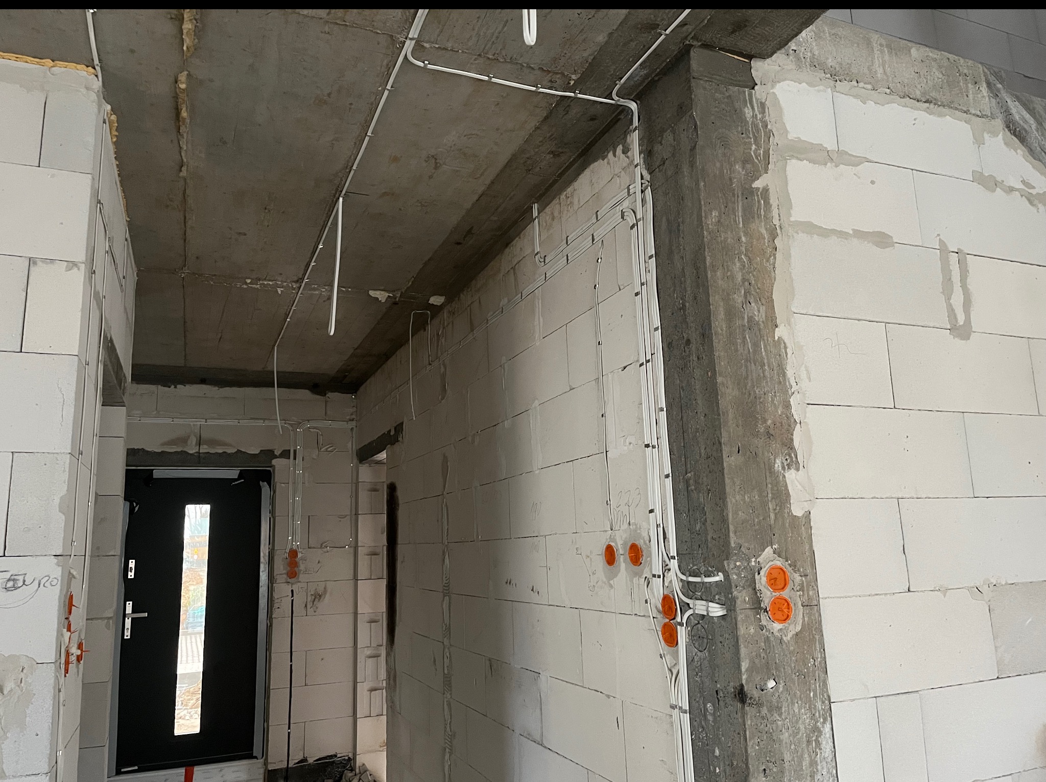 Surowy stan: białe ściany z betonu komórkowego, widoczne kable elektryczne w peszlach i pomarańczowe puszki podtynkowe. Instalacja w trakcie realizacji.