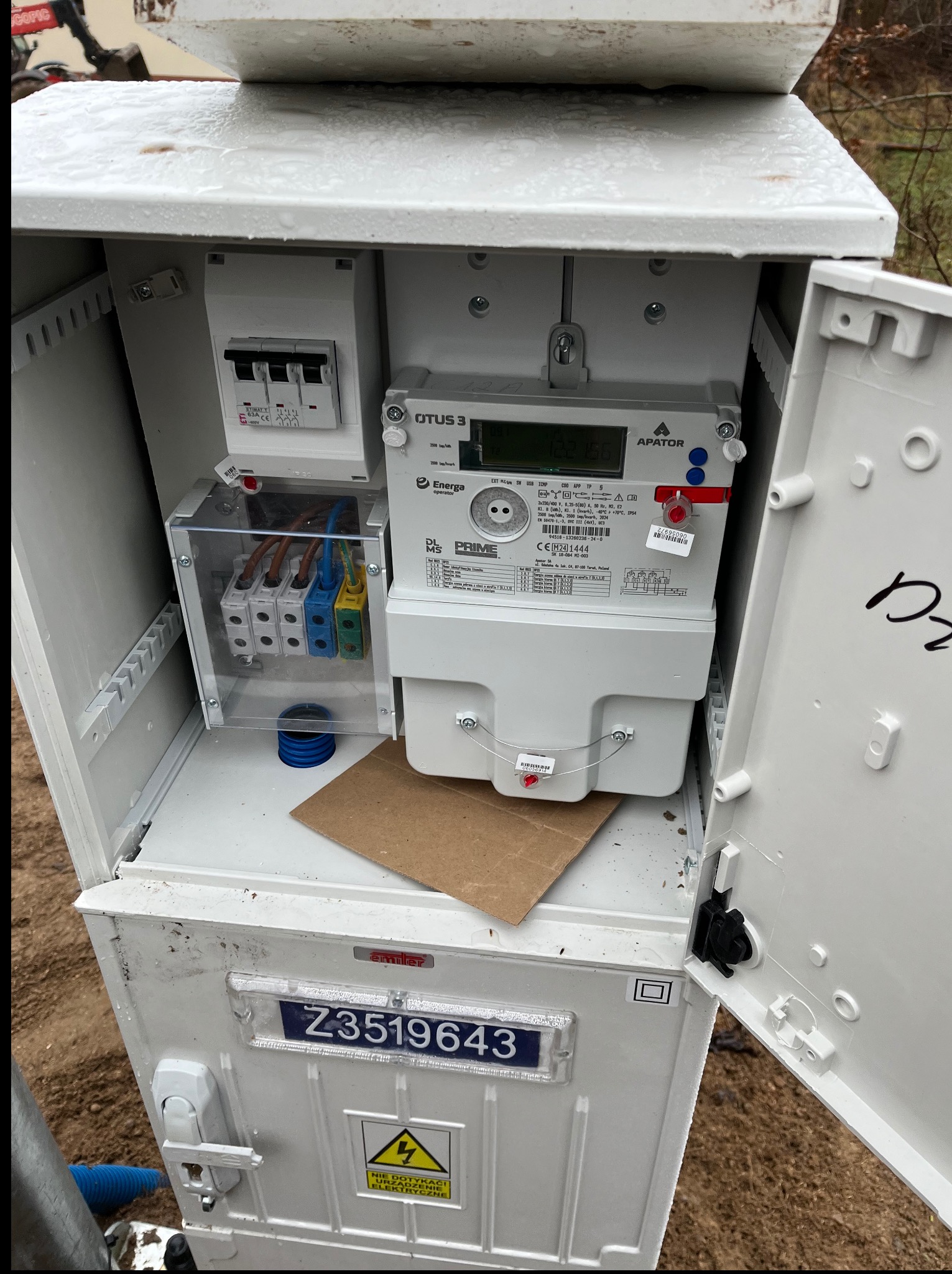 Otwarta skrzynka elektryczna z licznikiem Apator OTUS 3 i zabezpieczeniami, widoczne przewody i elementy instalacji. Tabliczka ostrzegawcza 'Nie dotykać urządzenia elektryczne'.