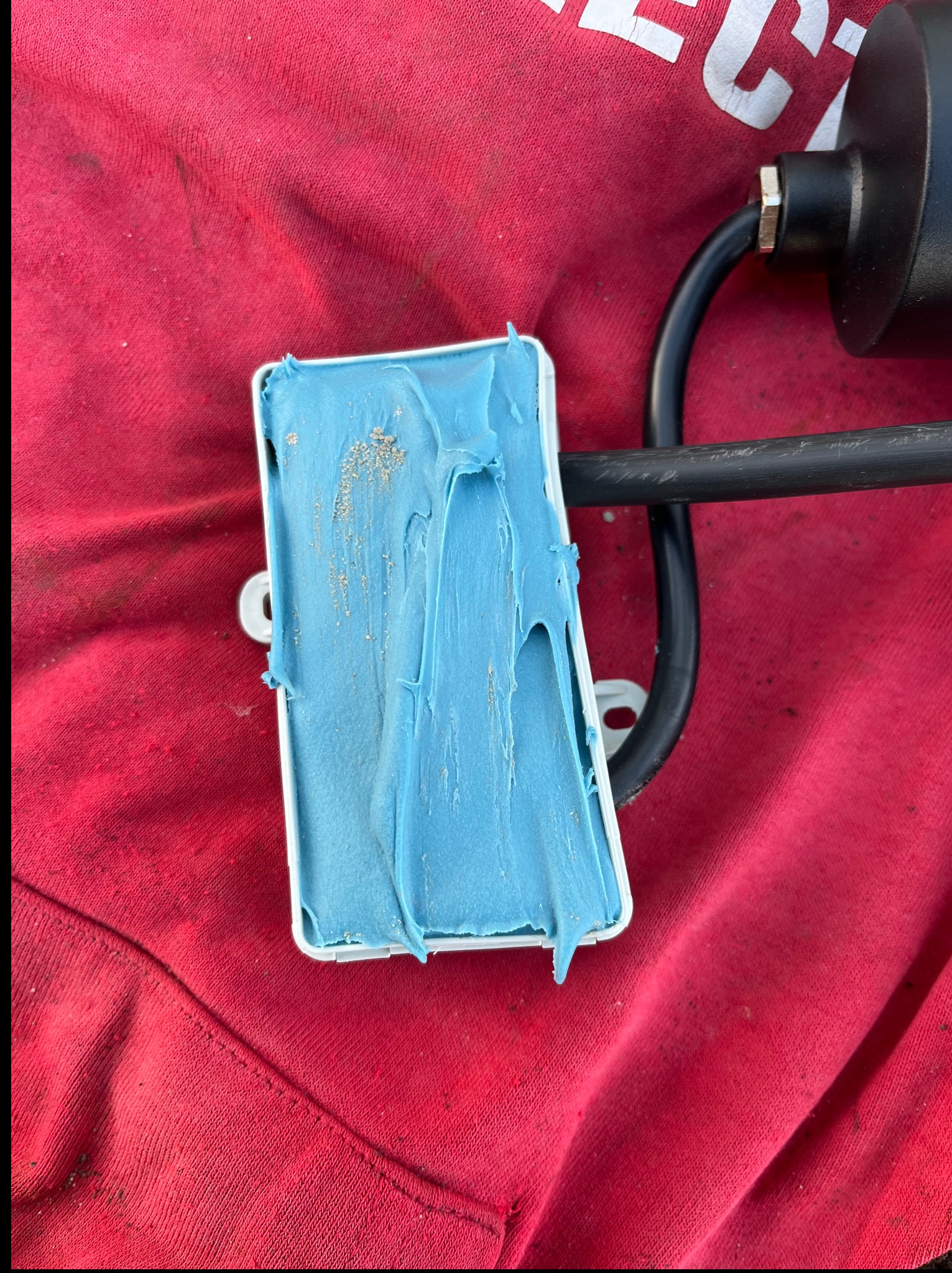 Uszczelniona puszka elektryczna z niebieską masą uszczelniającą, leżąca na czerwonej tkaninie. Widoczny fragment kabla i obudowy lampy.