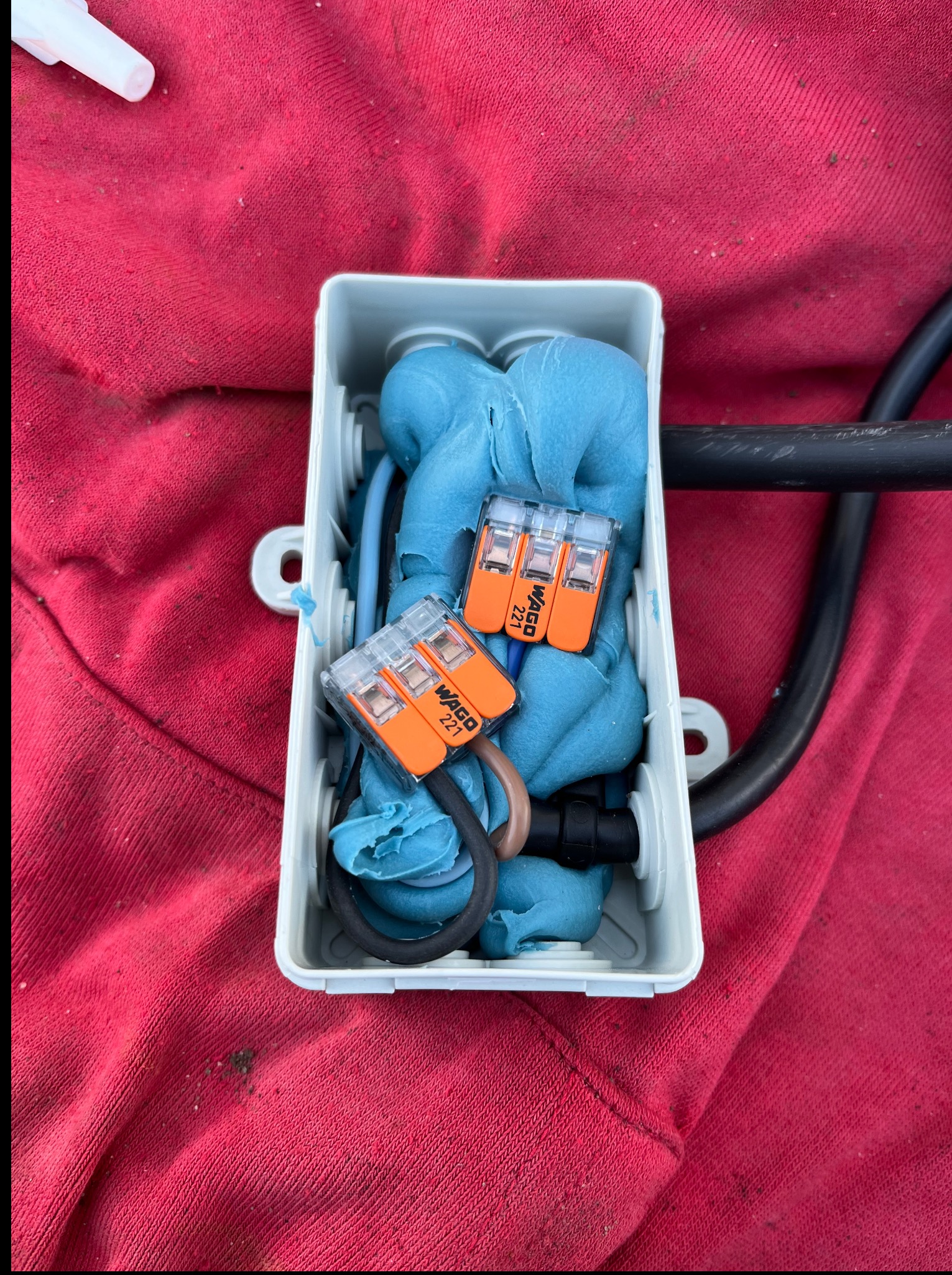 Otwarta puszka instalacyjna wypełniona niebieską masą uszczelniającą i złączkami WAGO 221, leżąca na czerwonym materiale. Widoczne przewody i fragment kabla.