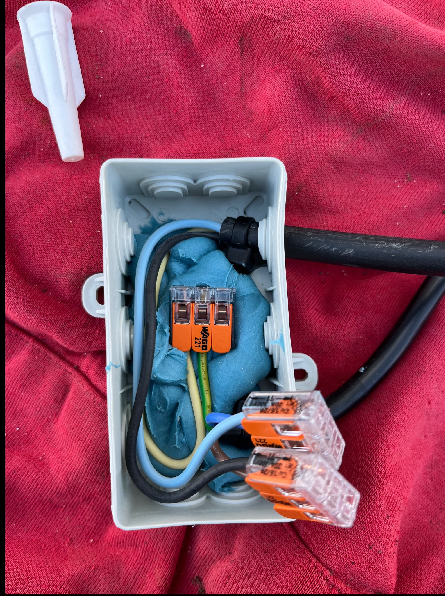 Szara puszka instalacyjna z przewodami i złączkami WAGO 221 na czerwonym tle. Widoczny fragment kabla i kołek rozporowy. Detal instalacji.