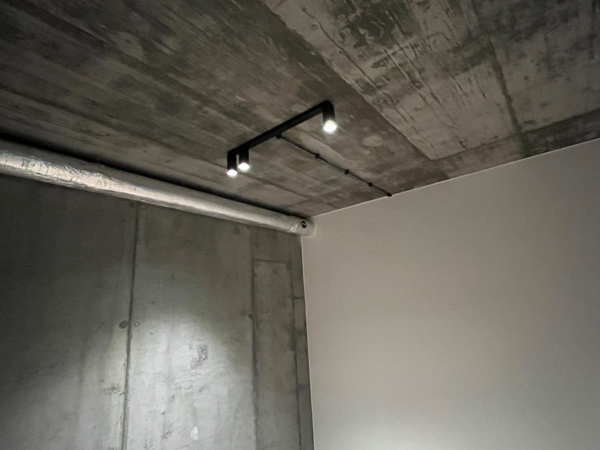 Minimalistyczna lampa sufitowa z trzema punktami świetlnymi na tle surowego, betonowego sufitu w nowoczesnym wnętrzu.