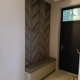 Nowoczesny przedpokój z tapicerowanym panelem ściennym w geometryczny wzór, złotymi haczykami i siedziskiem z szafką. Minimalistyczny design, stonowana kolorystyka.