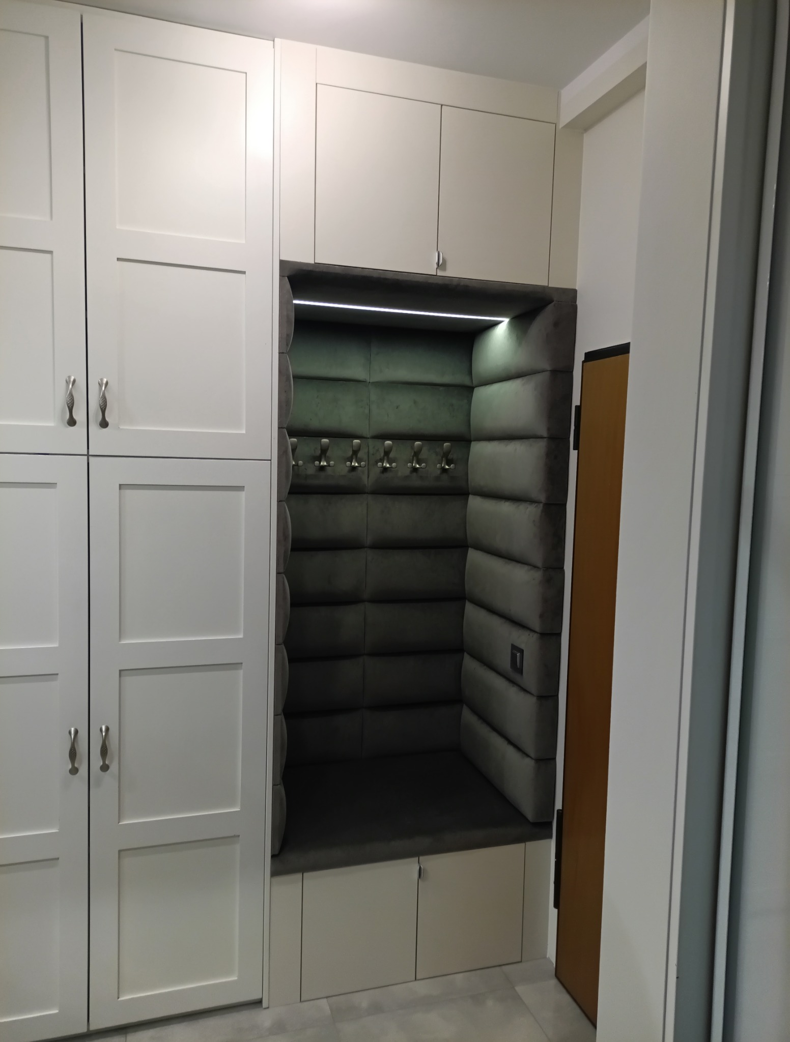 Biała szafa wnękowa z tapicerowanym siedziskiem i wieszakami, oświetlona LED, w nowoczesnym stylu. Szare, pikowane obicie dodaje elegancji.