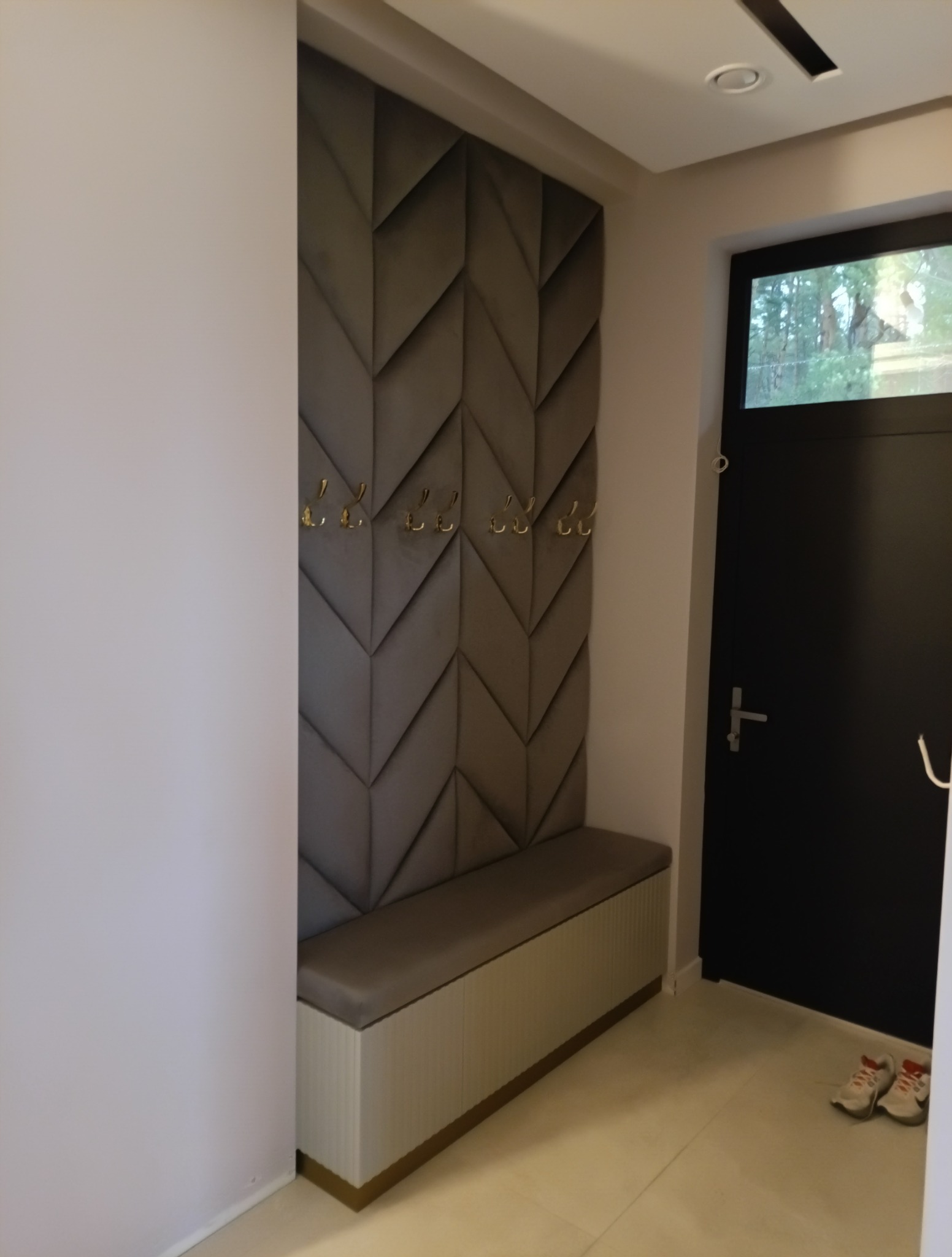 Nowoczesny przedpokój z tapicerowanym panelem ściennym w geometryczny wzór, złotymi haczykami i siedziskiem z szafką. Minimalistyczny design, stonowana kolorystyka.