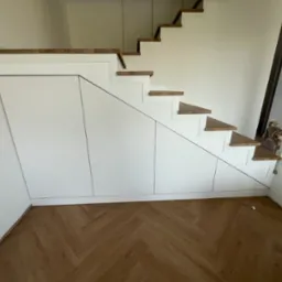 Białe szafy pod schodami z drewnianymi stopniami, parkiet w jodełkę na podłodze. Mały pies wchodzi po schodach, widoczny w tle.