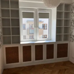 Biała zabudowa okienna z ażurowymi frontami szafek, otwartymi półkami i widokiem na budynek zza okna. Parkiet w jodełkę na podłodze.