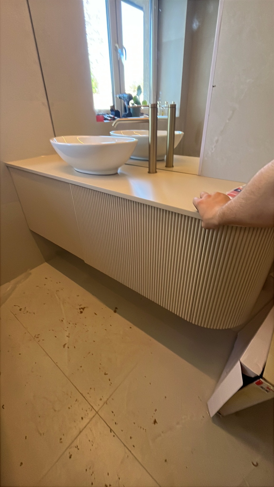 Beżowa szafka łazienkowa z umywalką nablatową i złotą armaturą, widoczna z boku, z elementami odbicia w lustrze i fragmentem dłoni w kadrze.