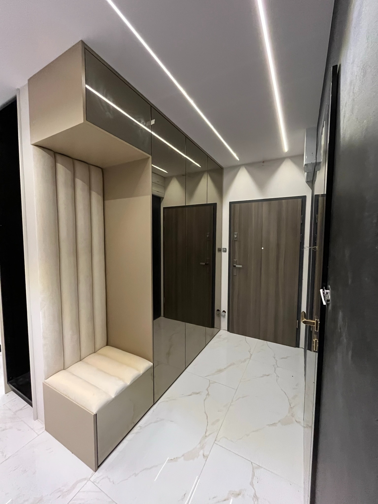 Nowoczesna szafa wnękowa z siedziskiem w przedpokoju, lustrzane fronty, oświetlenie LED, kremowa tapicerka, marmurowe płytki podłogowe, eleganckie wykończenie.
