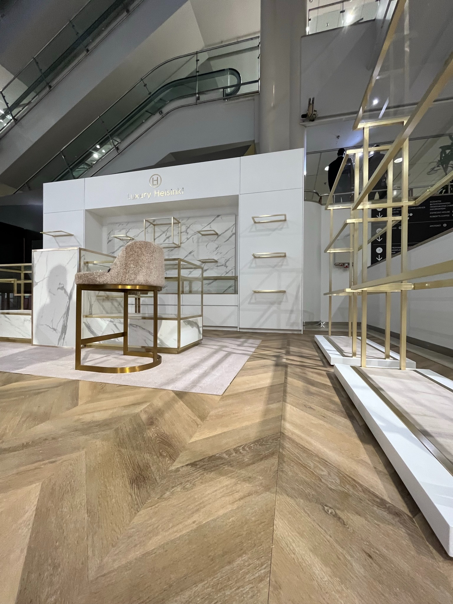 Biała zabudowa meblowa z marmurowym wzorem i złotymi akcentami, widoczna z perspektywy podłogi ułożonej w jodełkę, w tle ruchome schody.